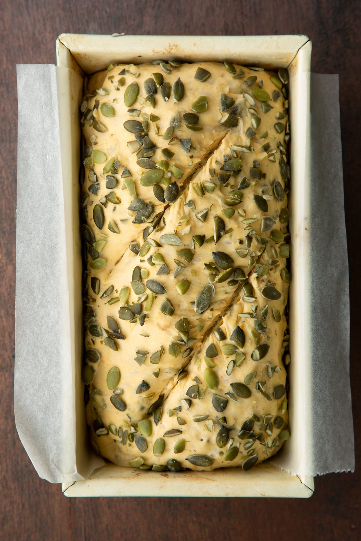 Proved pumpkin seed specie dough covered with pumpkin seeds in a loaf tin lined with sultry paper. The top has been slashed twice.