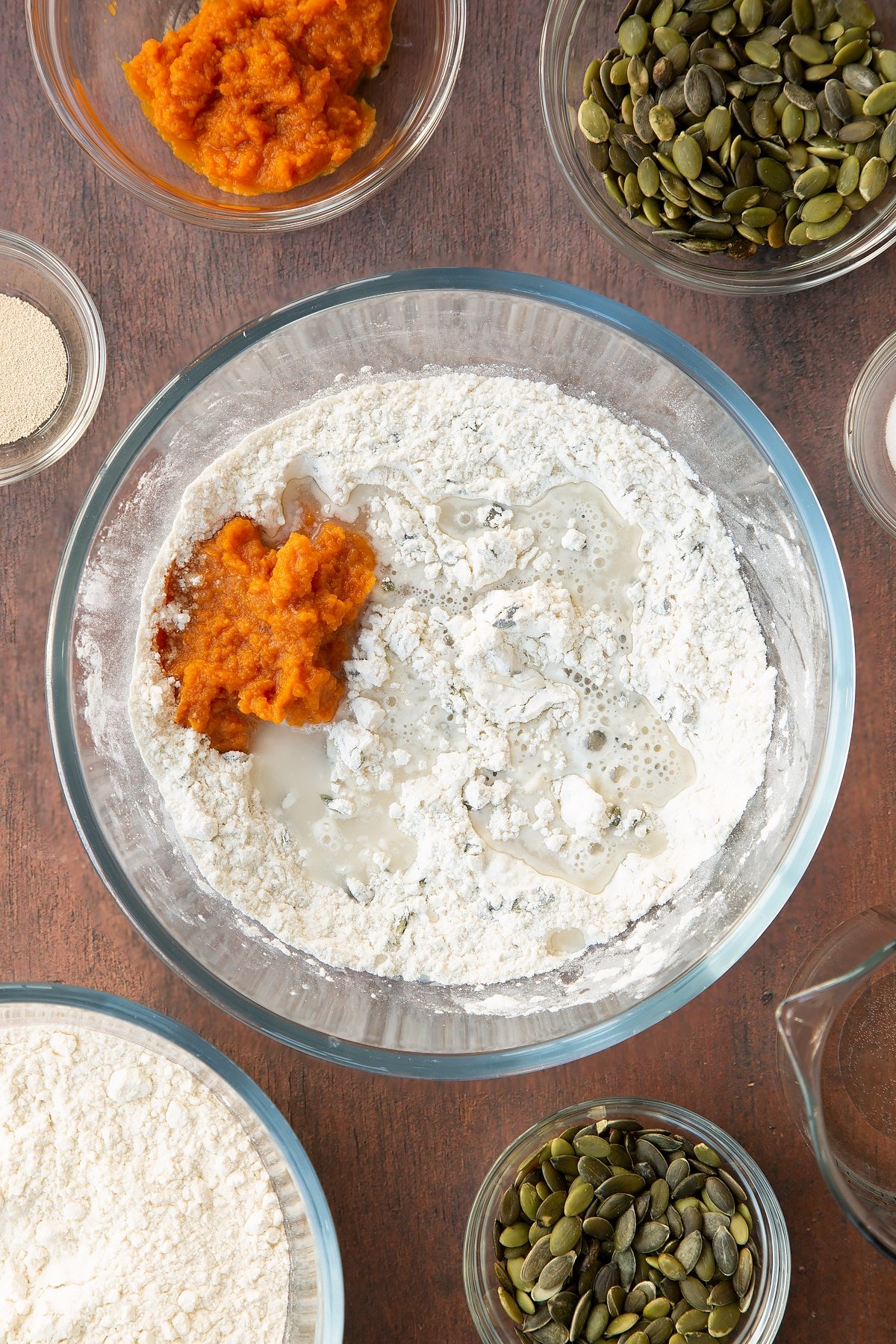 Flour, salt, yeast and pumpkin seeds mixed together in a glass mixing trencher with pumpkin puree and water on top. Ingredients to make a pumpkin seed specie recipe surround the bowl. 