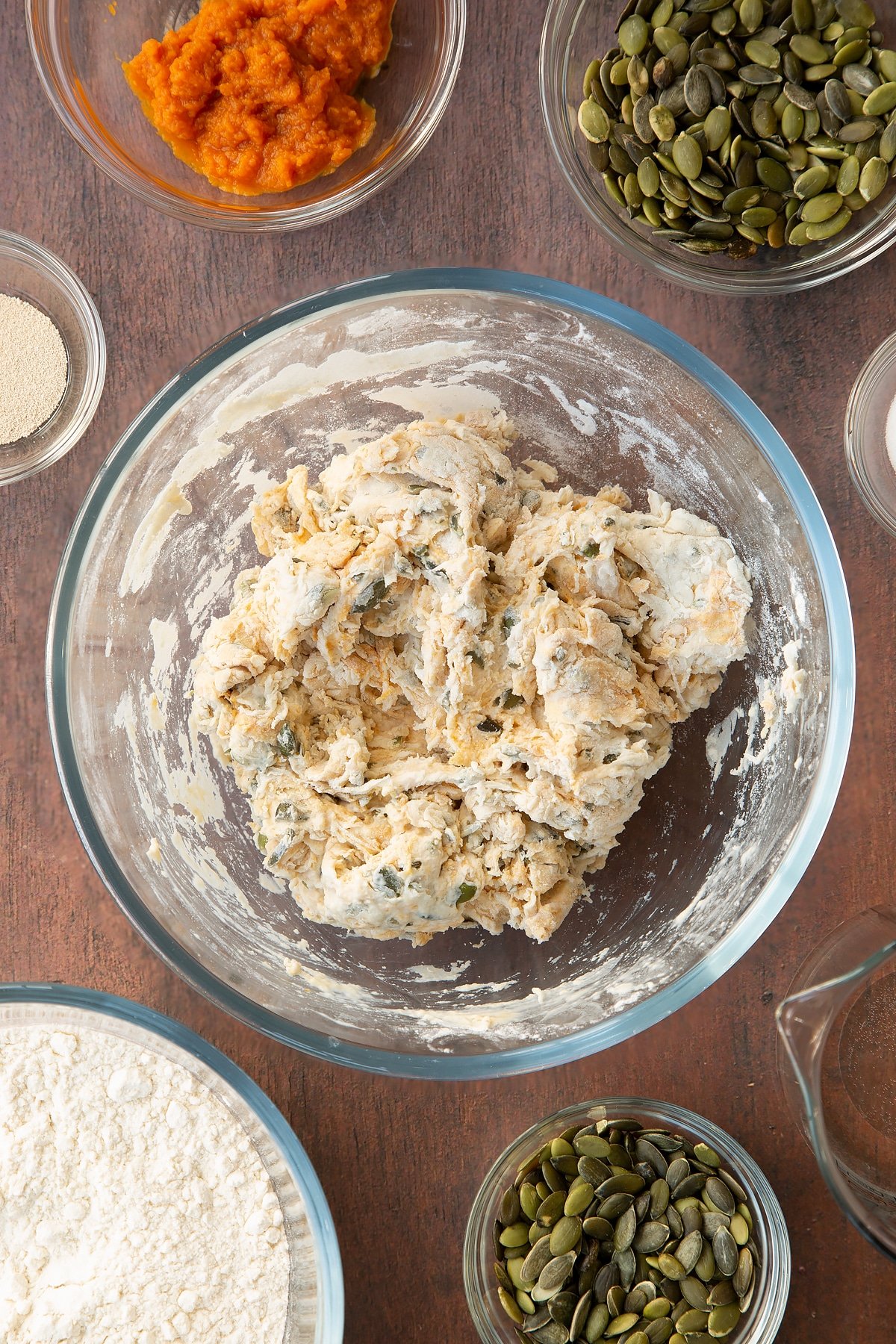 Rough pumpkin seed specie dough in a glass mixing bowl. Ingredients to make a pumpkin seed specie recipe surround the bowl. 