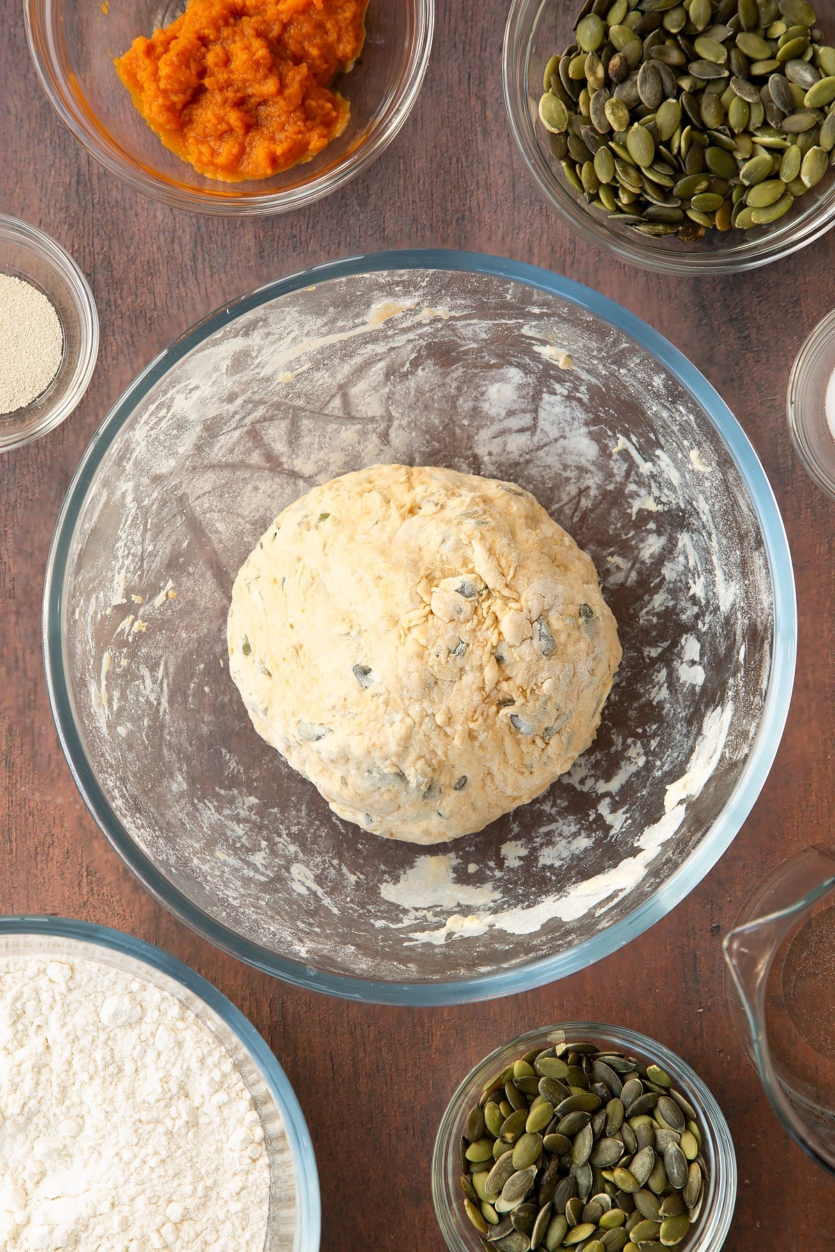 A ball of pumpkin seed bread dough in a glass mixing bowl. Ingredients to make a pumpkin seed bread recipe surround the bowl. 