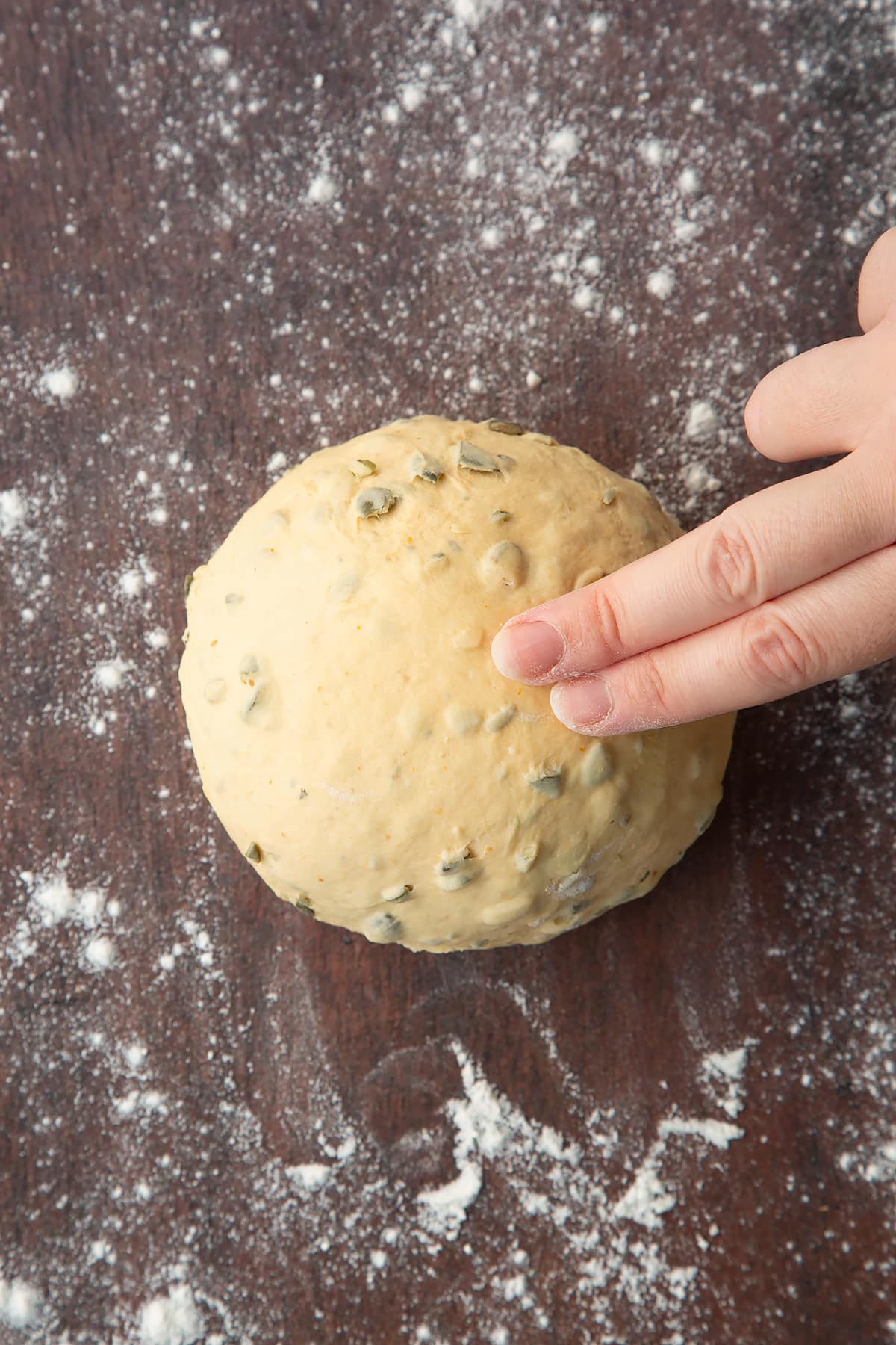 Two fingers pressing a ball of pumpkin seed bread dough on a floured surface.