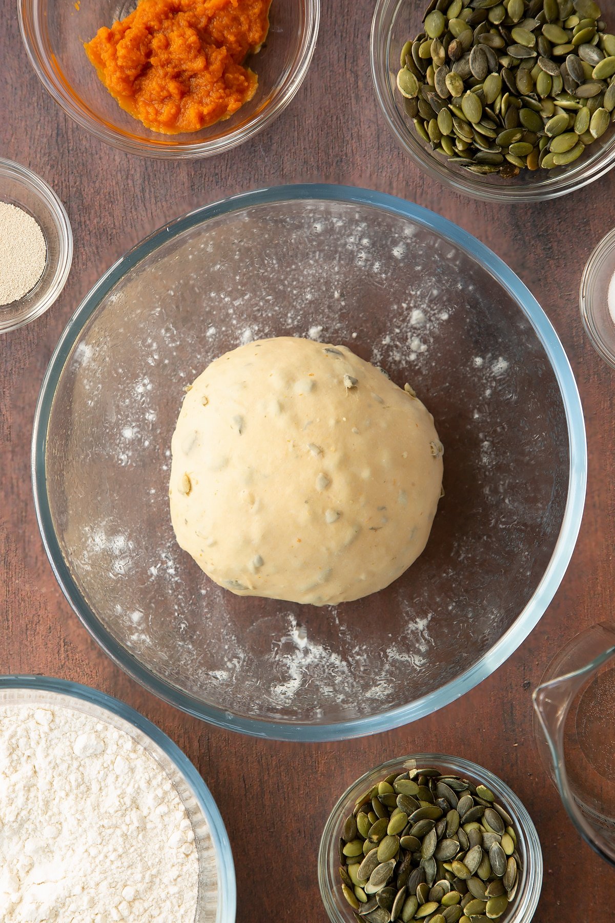 A ball of kneaded pumpkin seed bread dough in a glass mixing bowl. Ingredients to make a pumpkin seed bread recipe surround the bowl. 