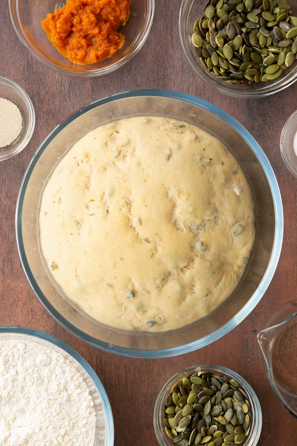 A ball of proved pumpkin seed bread dough in a glass mixing bowl. Ingredients to make a pumpkin seed bread recipe surround the bowl. 