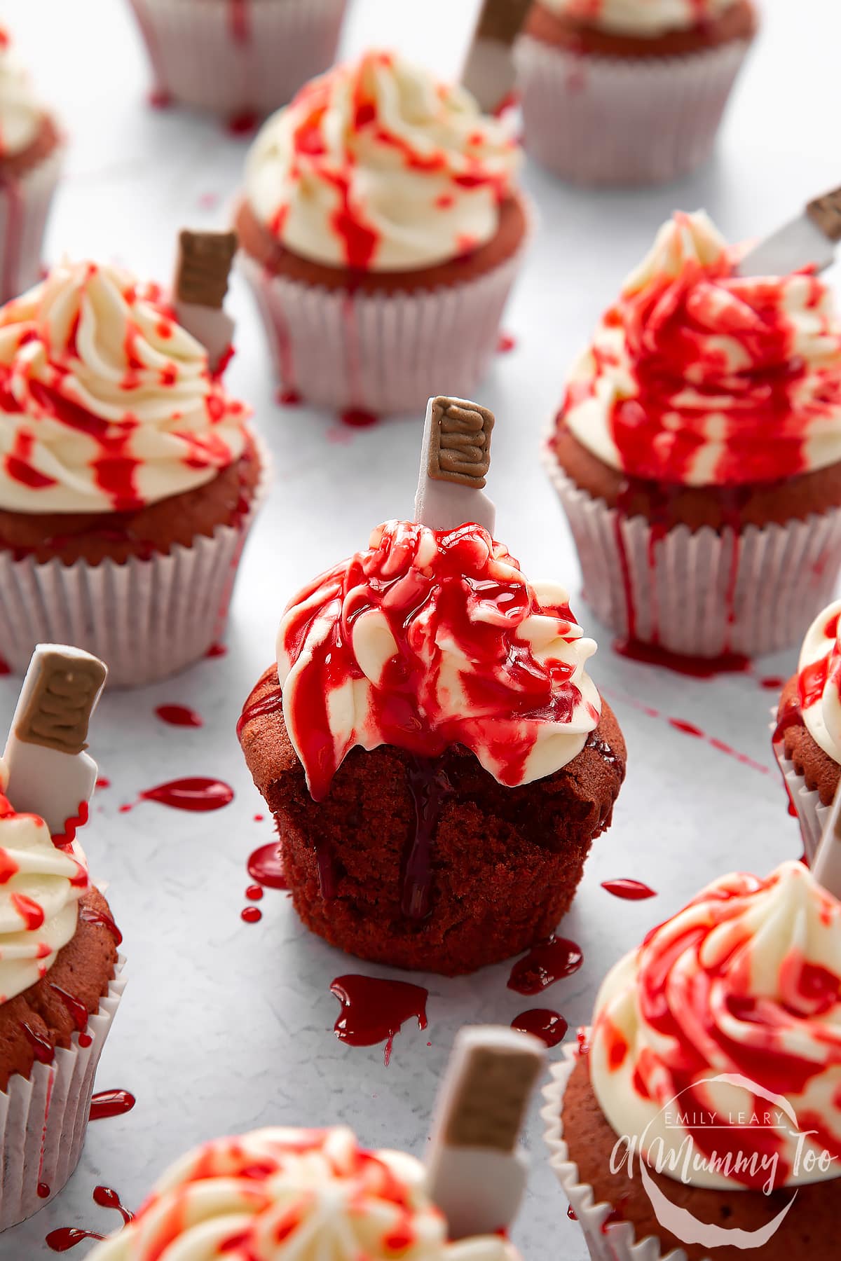 Red velvet Halloween cupcakes, spattered with red syrup. The one in the foreground has been unwrapped and has a bite out of it.