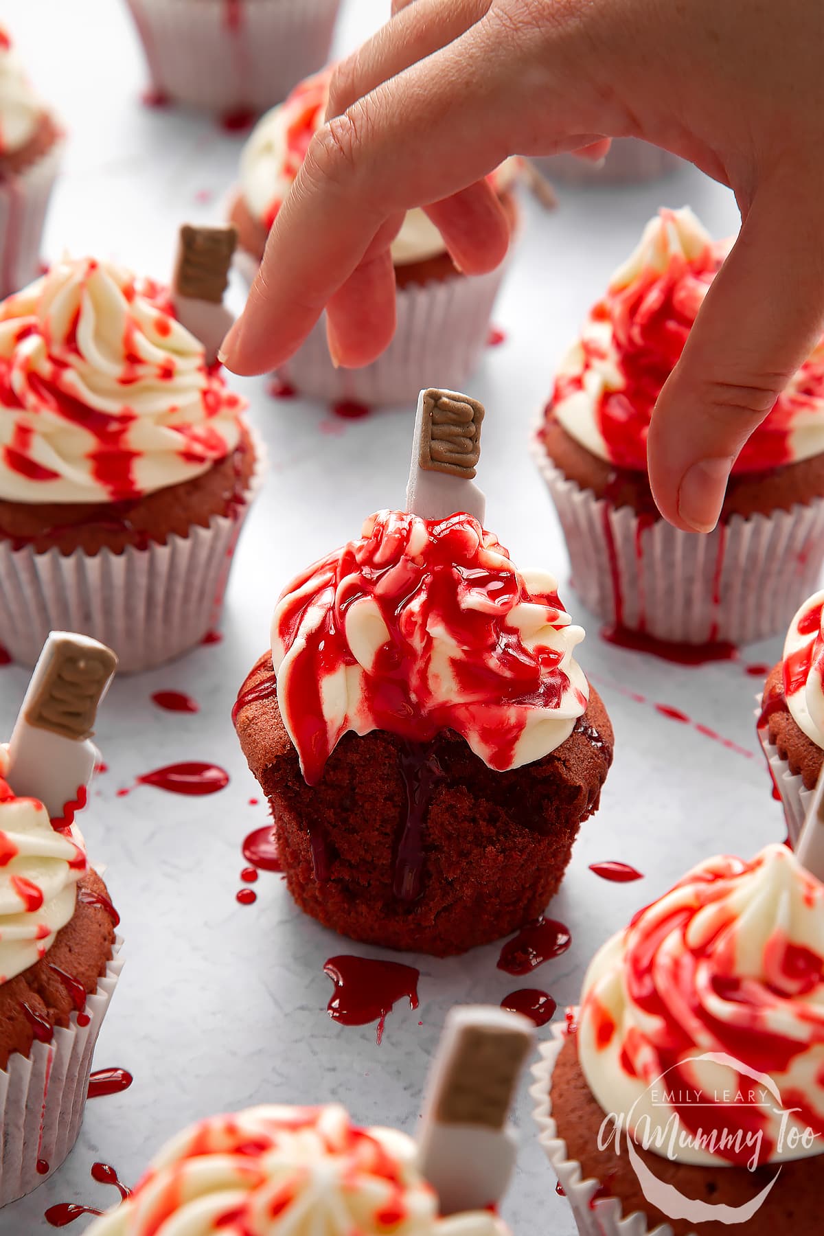 Red velvet Halloween cupcakes, spattered with red syrup. A hand reaches for one with a bite out of it.