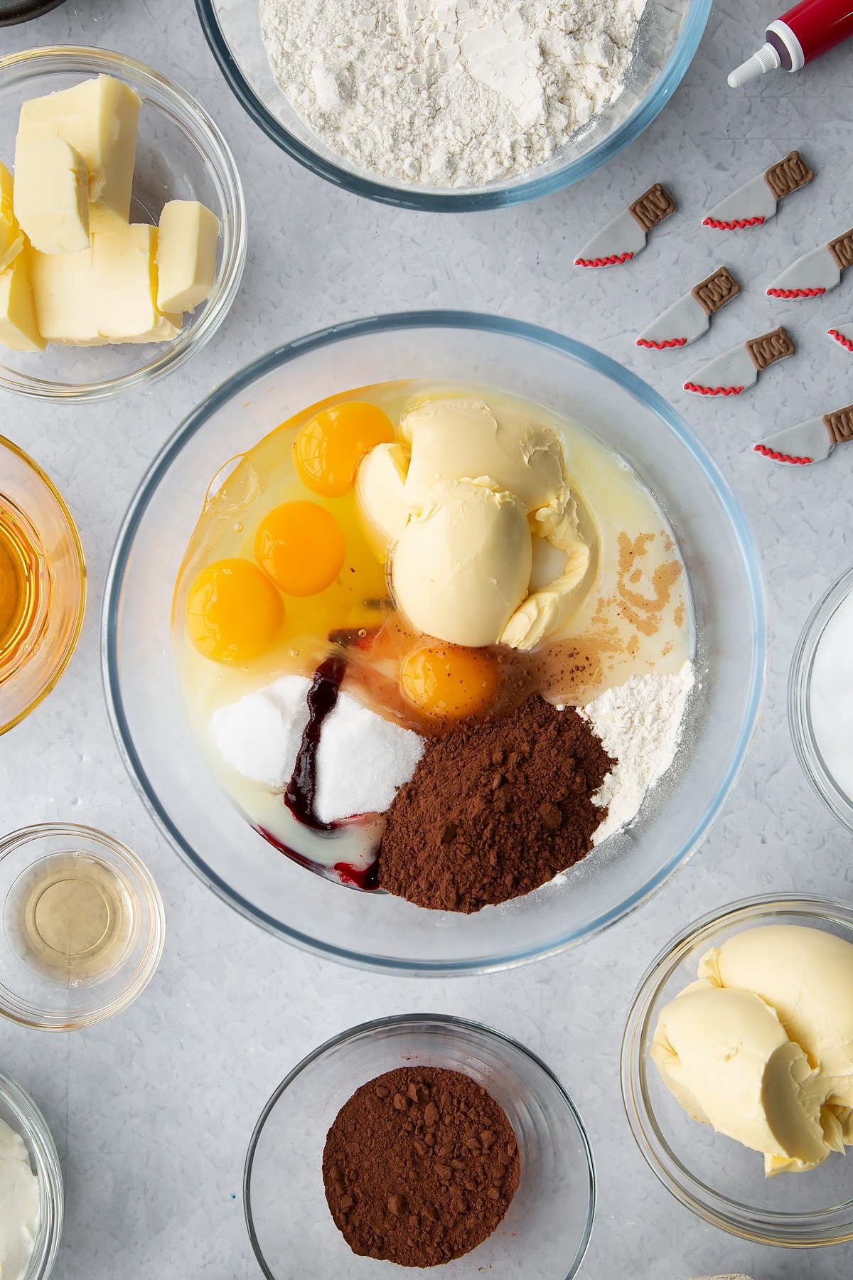 Margarine, milk, sugar, flour, cocoa, eggs and food colouring in a glass bowl. Ingredients to make red velvet Halloween cupcakes surround the bowl. 