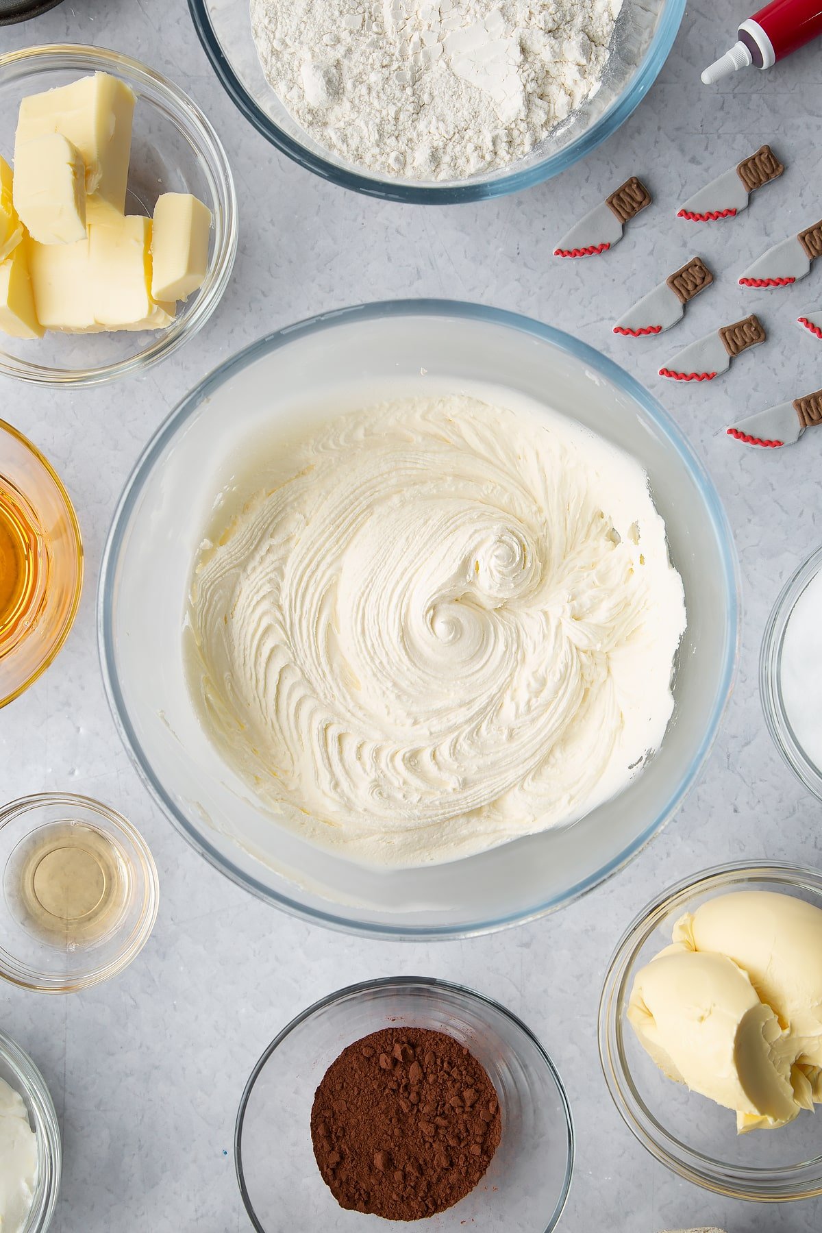Vanilla buttercream in a glass bowl. Ingredients to make red velvet Halloween cupcakes surround the bowl.