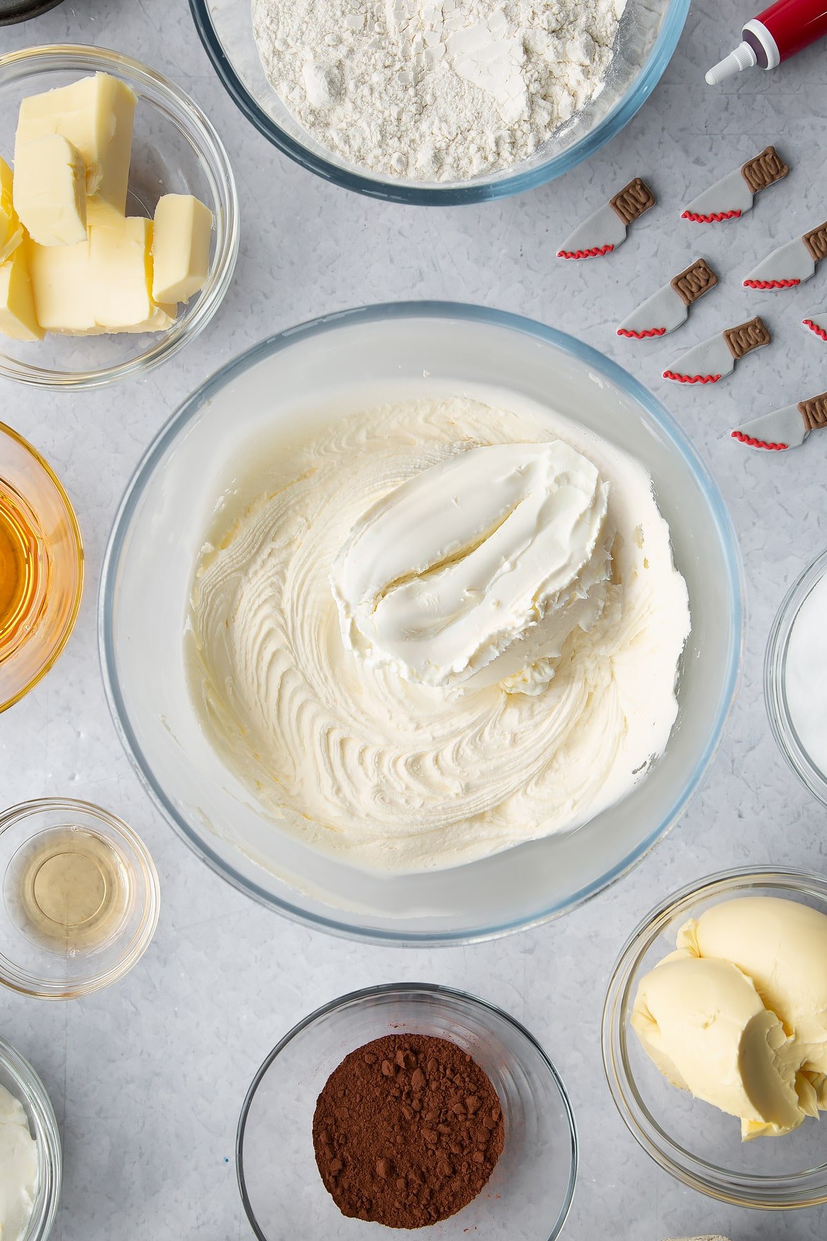 Vanilla buttercream in a glass bowl with cream cheese on top. Ingredients to make red velvet Halloween cupcakes surround the bowl.