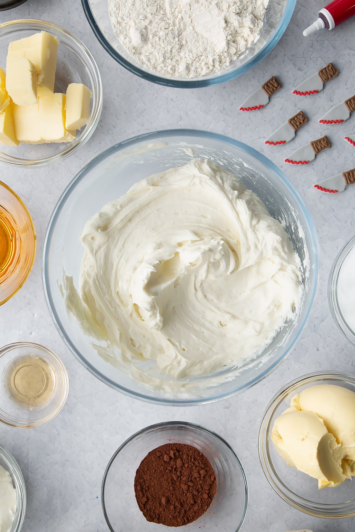 Cream cheese frosting in a glass bowl. Ingredients to make red velvet Halloween cupcakes surround the bowl. 