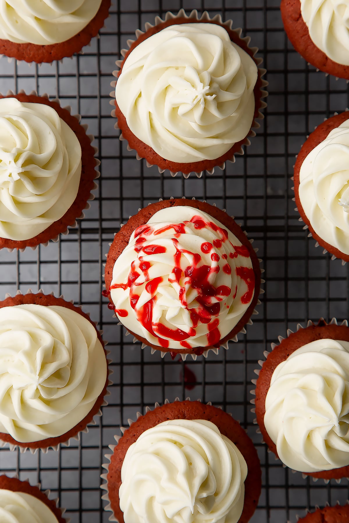 Cream cheese frosting piped on top of red velvet Halloween cupcakes on a wire rack. One has been drizzled with red syrup.