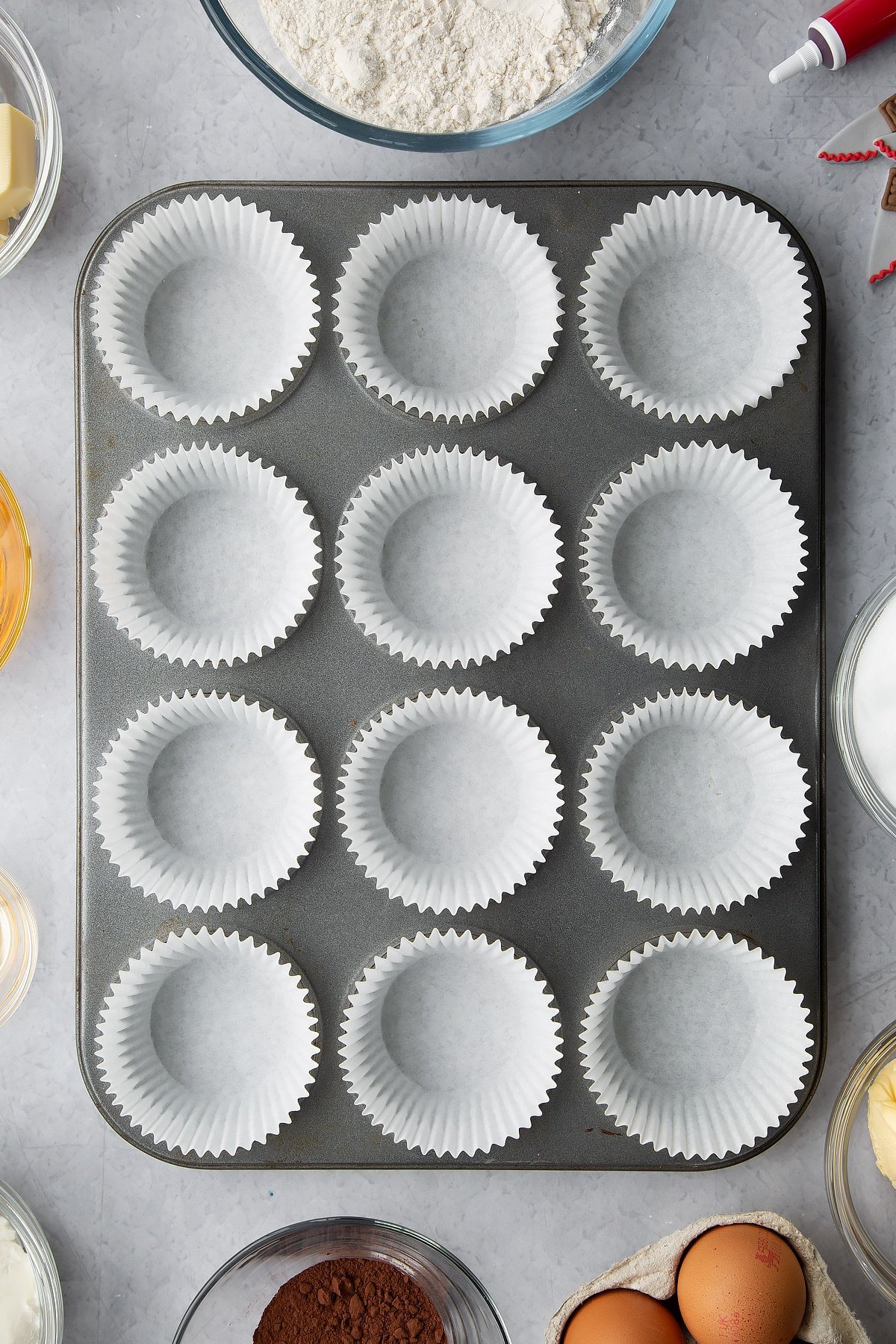 A muffin tray lined with cupcake cases. Ingredients to make red velvet Halloween cupcakes surround the tin. 