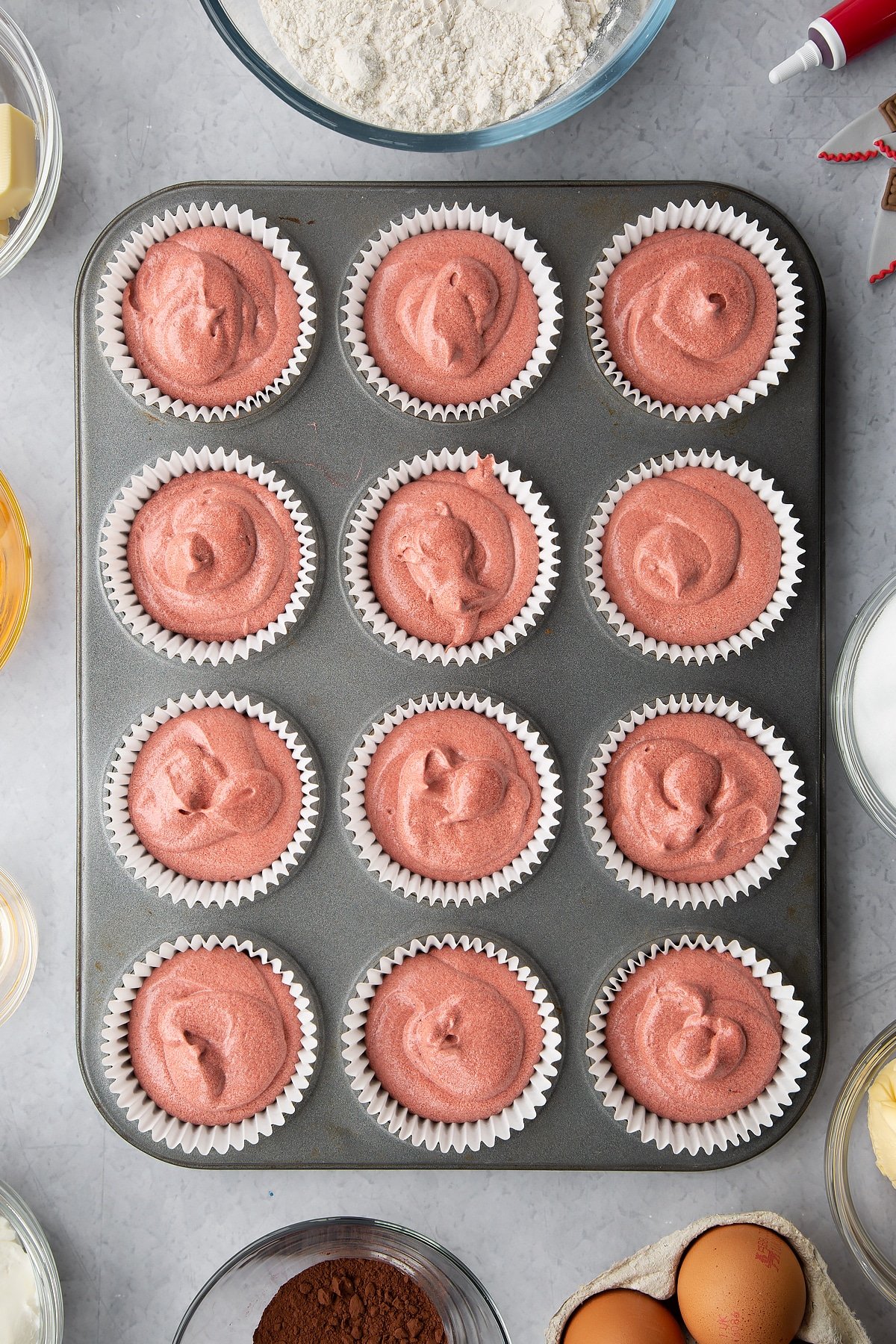 Red velvet cake batter in a muffin tray lined with cupcake cases. Ingredients to make red velvet Halloween cupcakes surround the tin. 