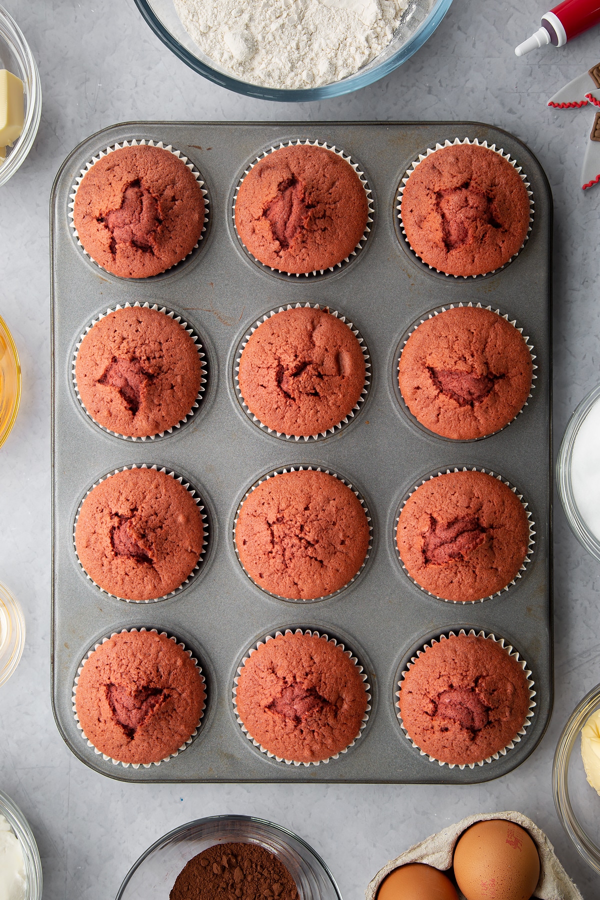 Red velvet cupcakes in a muffin tray. Ingredients to make red velvet Halloween cupcakes surround the tin.