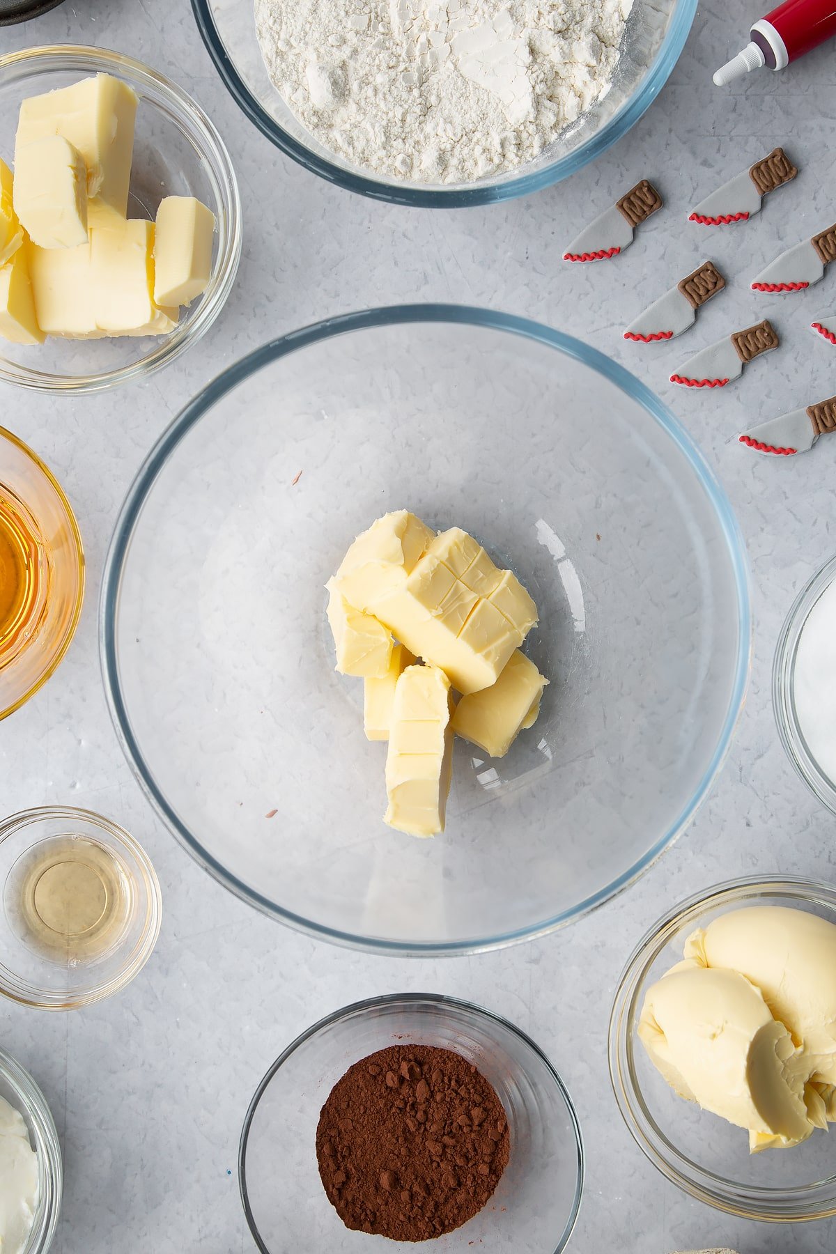 Cubed butter in a glass bowl. Ingredients to make red velvet Halloween cupcakes surround the bowl. 