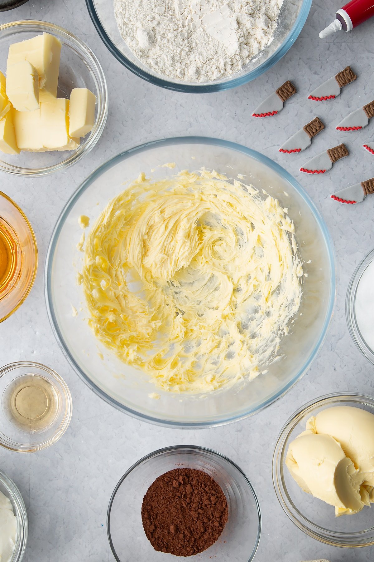 Whipped butter in a glass bowl. Ingredients to make red velvet Halloween cupcakes surround the bowl. 