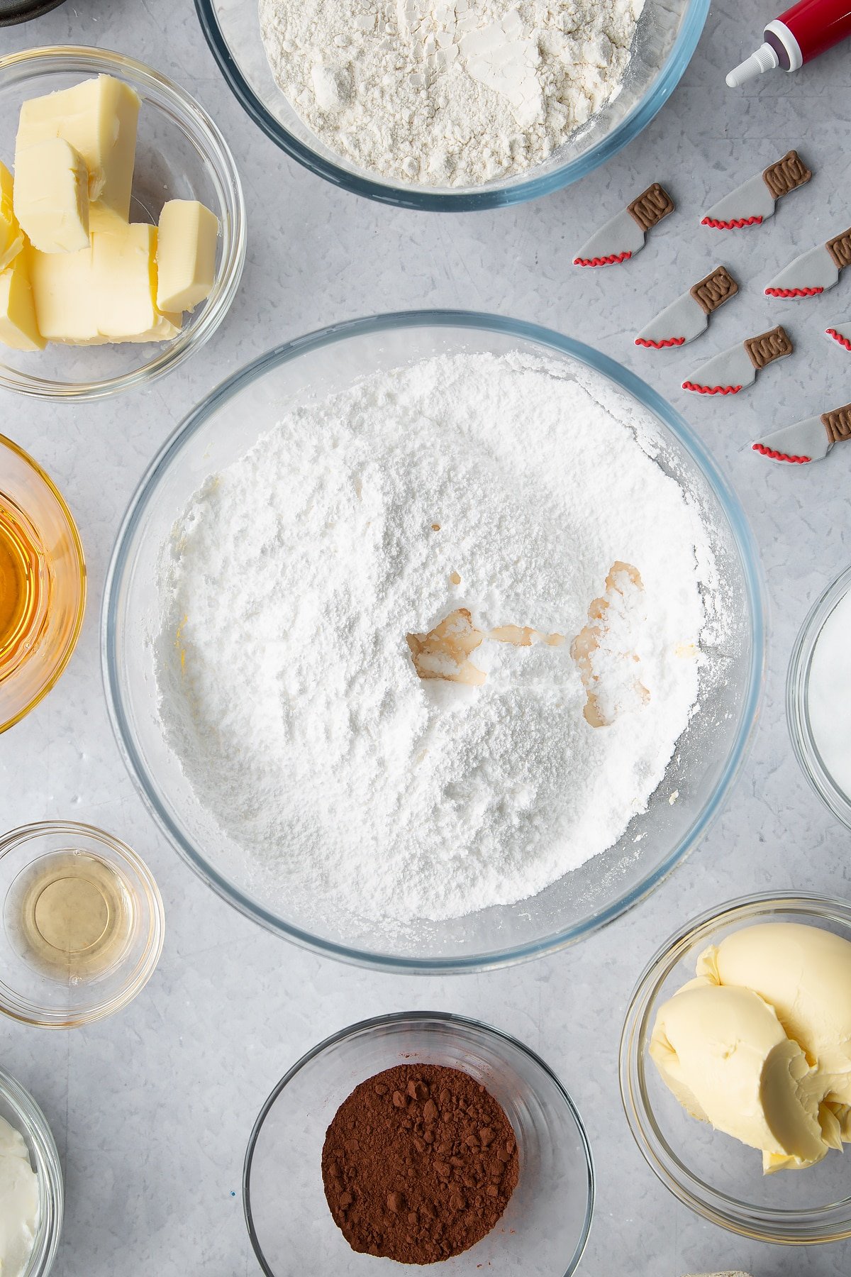 Butter, icing sugar and vanilla extract in a glass bowl. Ingredients to make red velvet Halloween cupcakes surround the bowl.