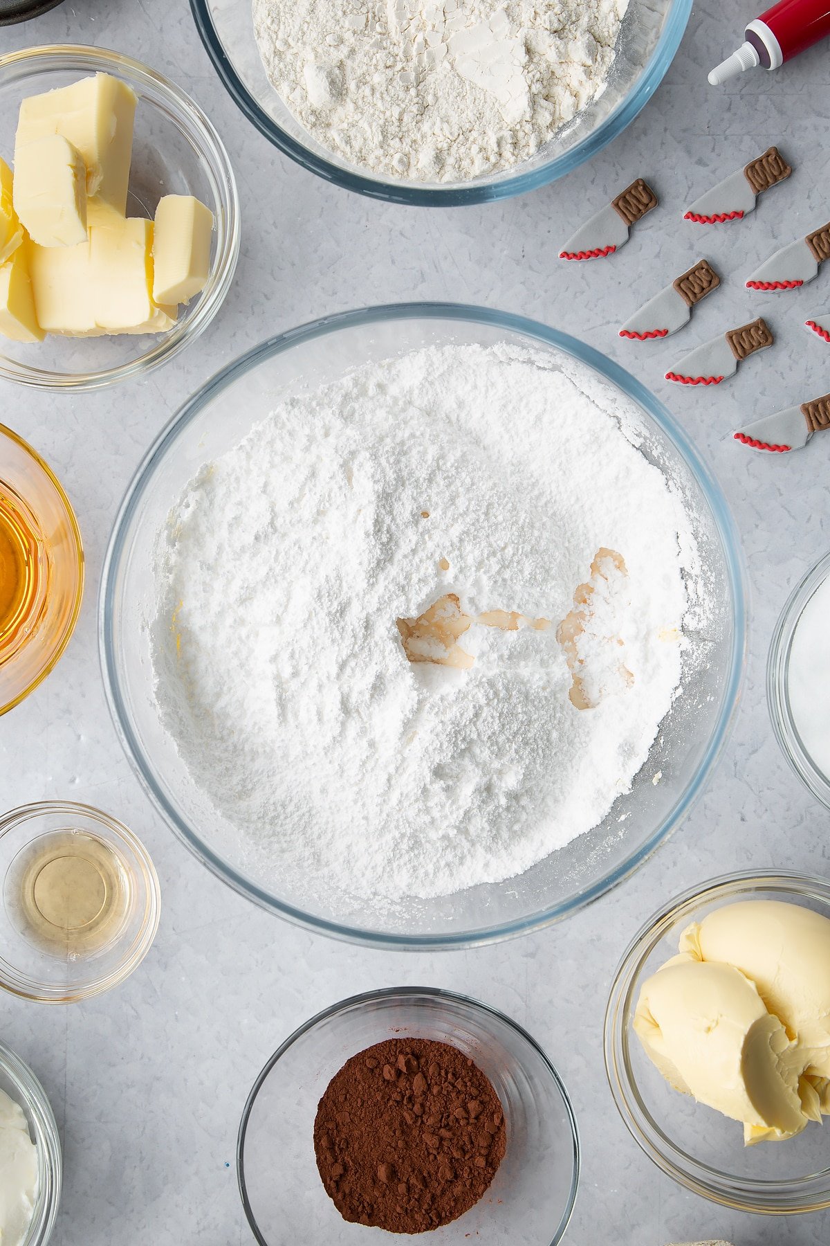 Butter, icing sugar and vanilla extract in a glass bowl. Ingredients to make red velvet Halloween cupcakes surround the bowl. 