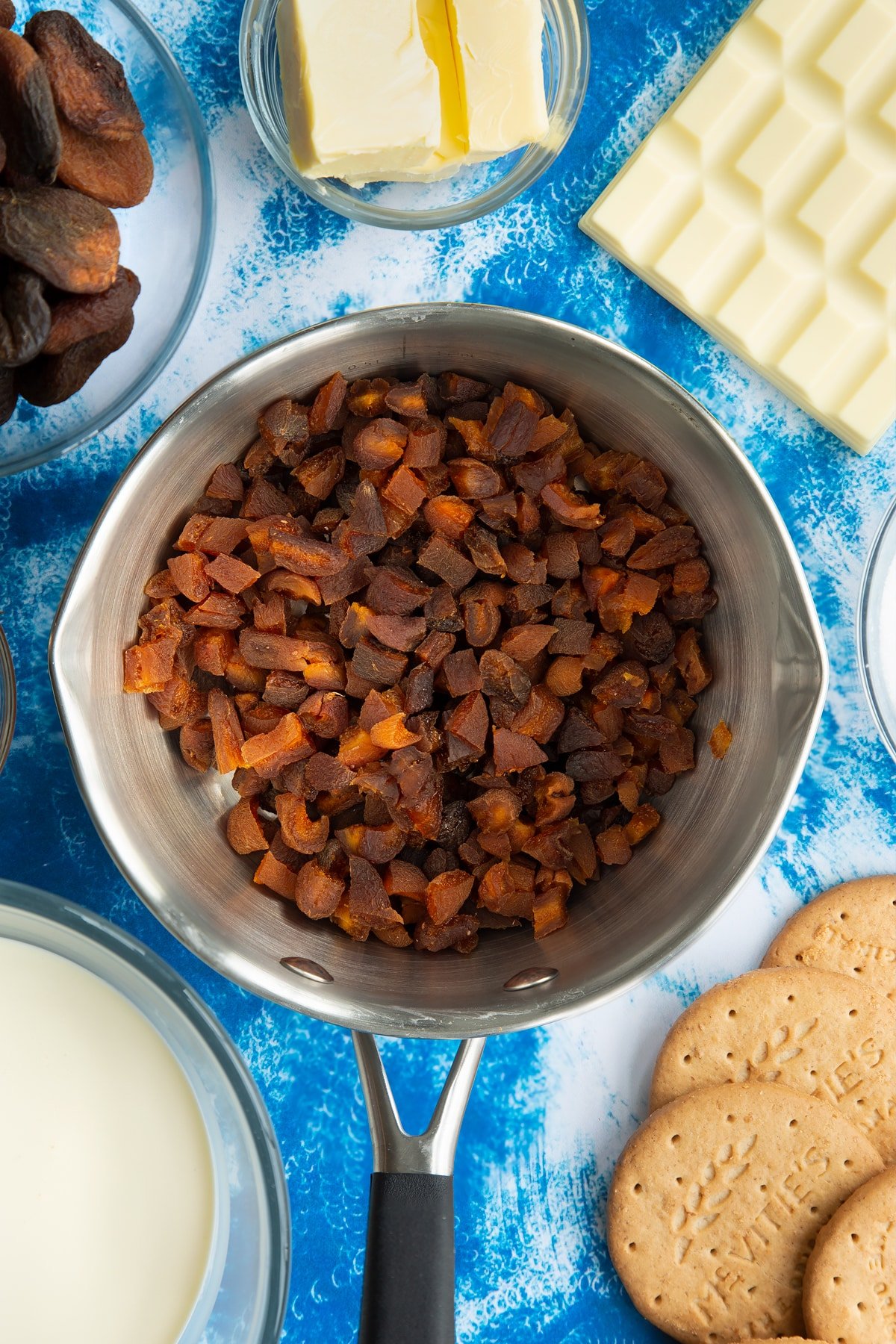 Chopped dried apricots in a pan. Ingredients to make an apricot cheesecake surround the pan.