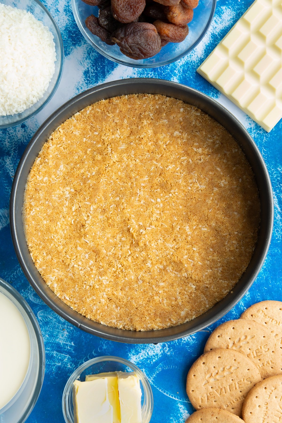 Crushed biscuits, coconut and butter pressed into the base of a tin. Ingredients to make an apricot cheesecake surround the tin.