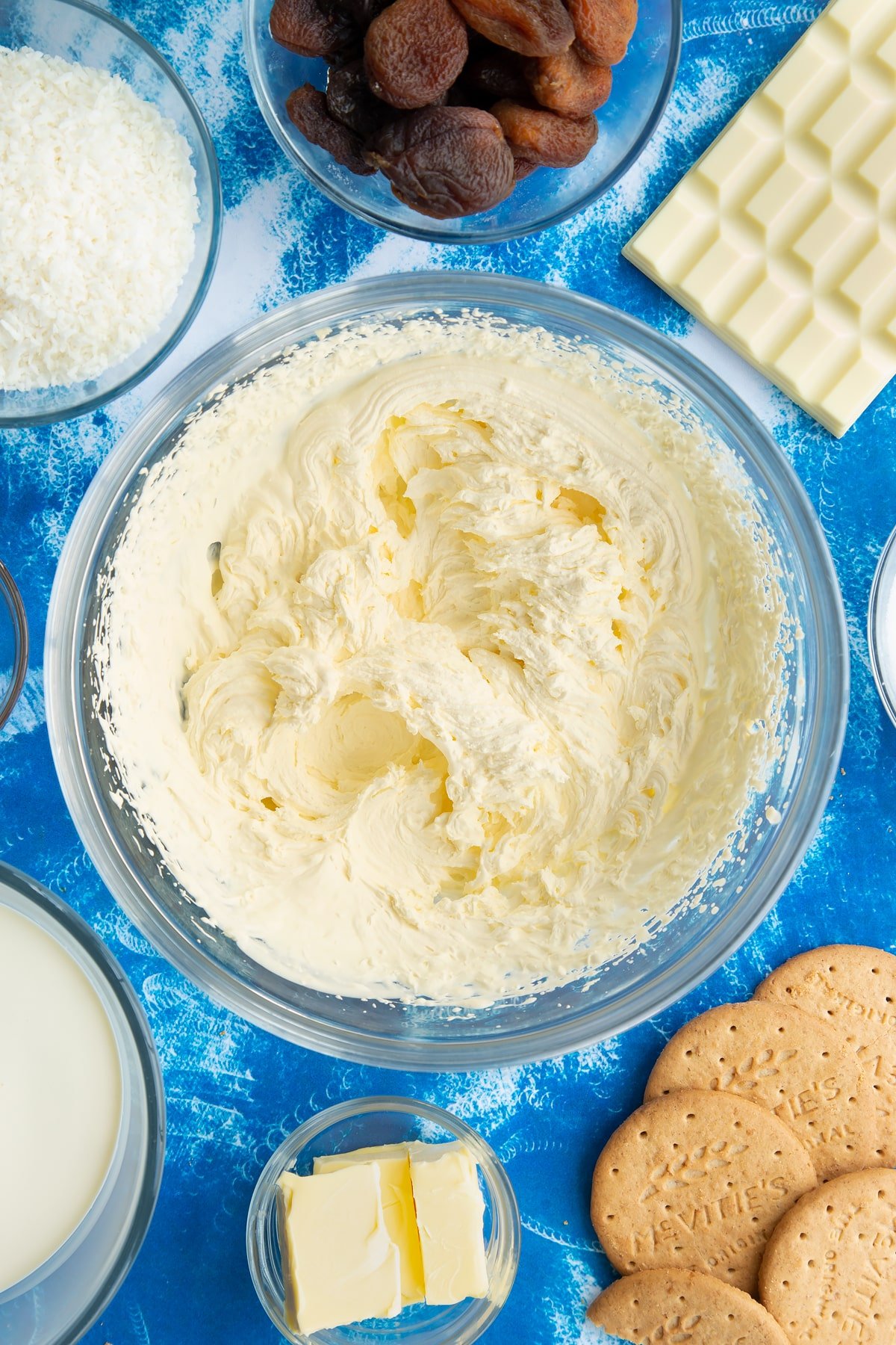 Whipped double cream in a bowl. Ingredients to make an apricot cheesecake surround the bowl.