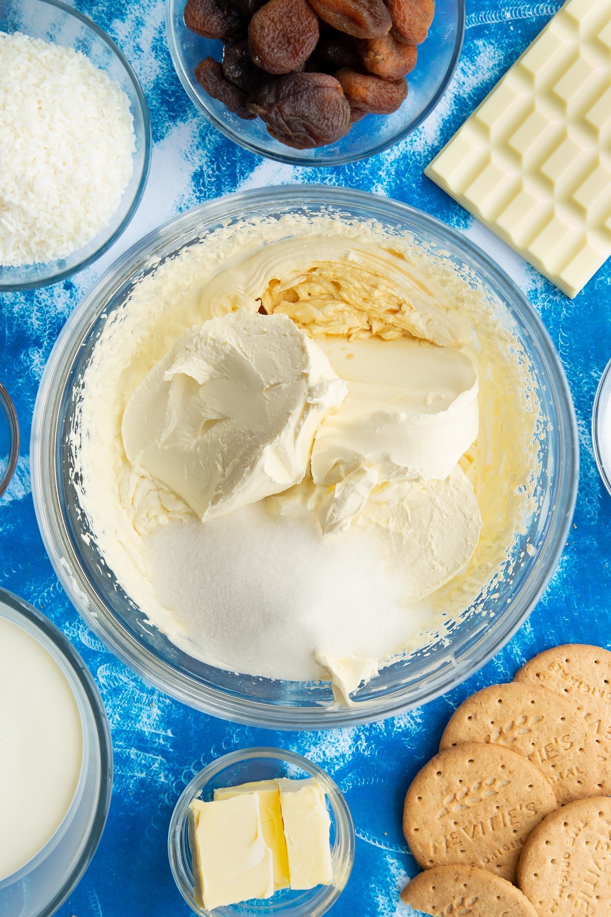 Whipped double cream with cream cheese, vanilla and sugar on top in a bowl. Ingredients to make an apricot cheesecake surround the bowl.