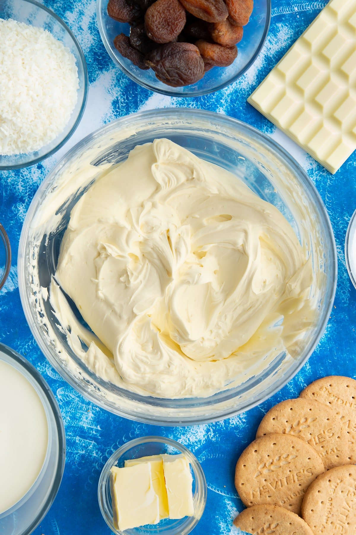 Whipped double cream, cream cheese, vanilla and sugar folded together in a bowl. Ingredients to make an apricot cheesecake surround the bowl.