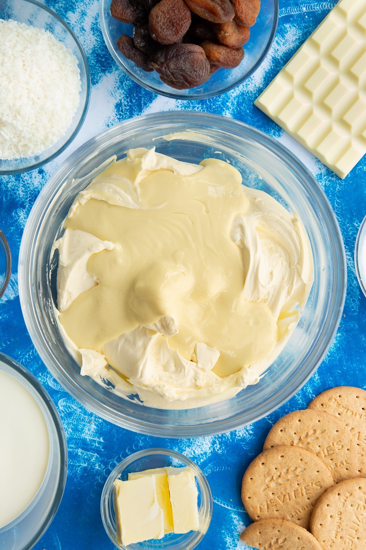 Whipped double cream, cream cheese, vanilla and sugar folded together in a bowl with melted white chocolate on top. Ingredients to make an apricot cheesecake surround the bowl.