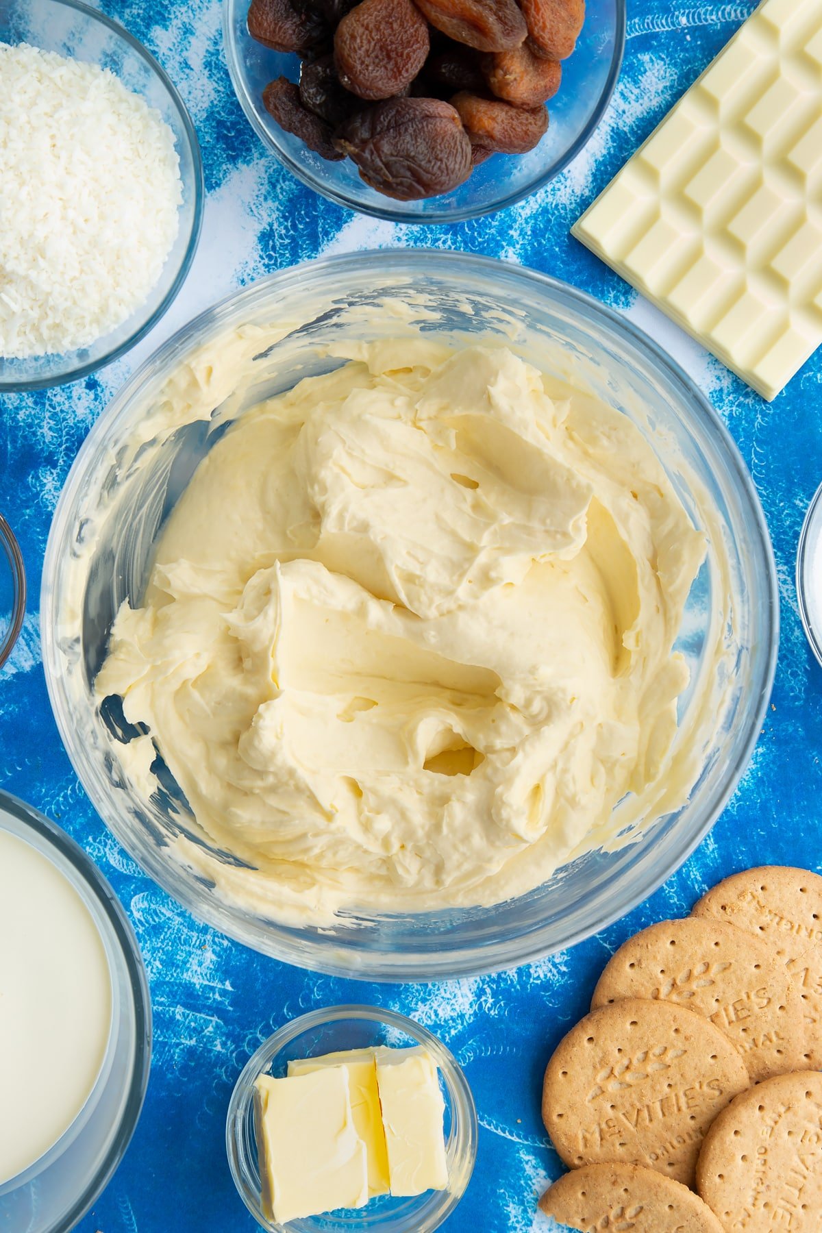 Whipped double cream, cream cheese, vanilla, sugar and melted white chocolate folded together in a bowl. Ingredients to make an apricot cheesecake surround the bowl.