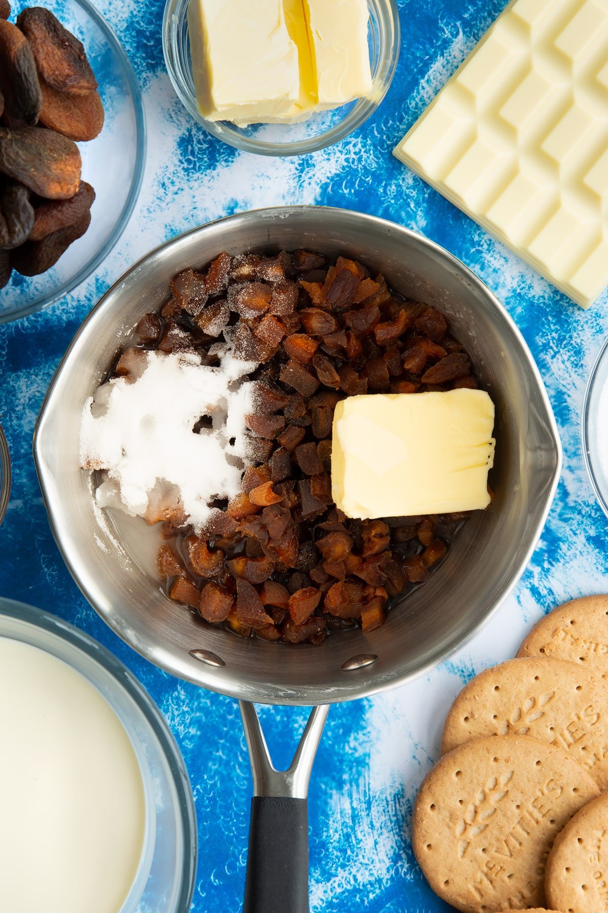 Chopped dried apricots, sugar, butter and water in a pan. Ingredients to make an apricot cheesecake surround the pan.