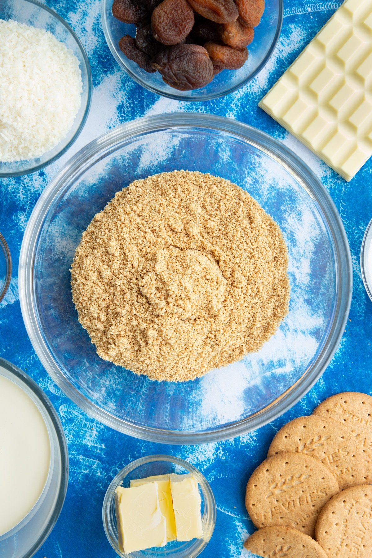 Crushed biscuits in a bowl. Ingredients to make an apricot cheesecake surround the bowl.