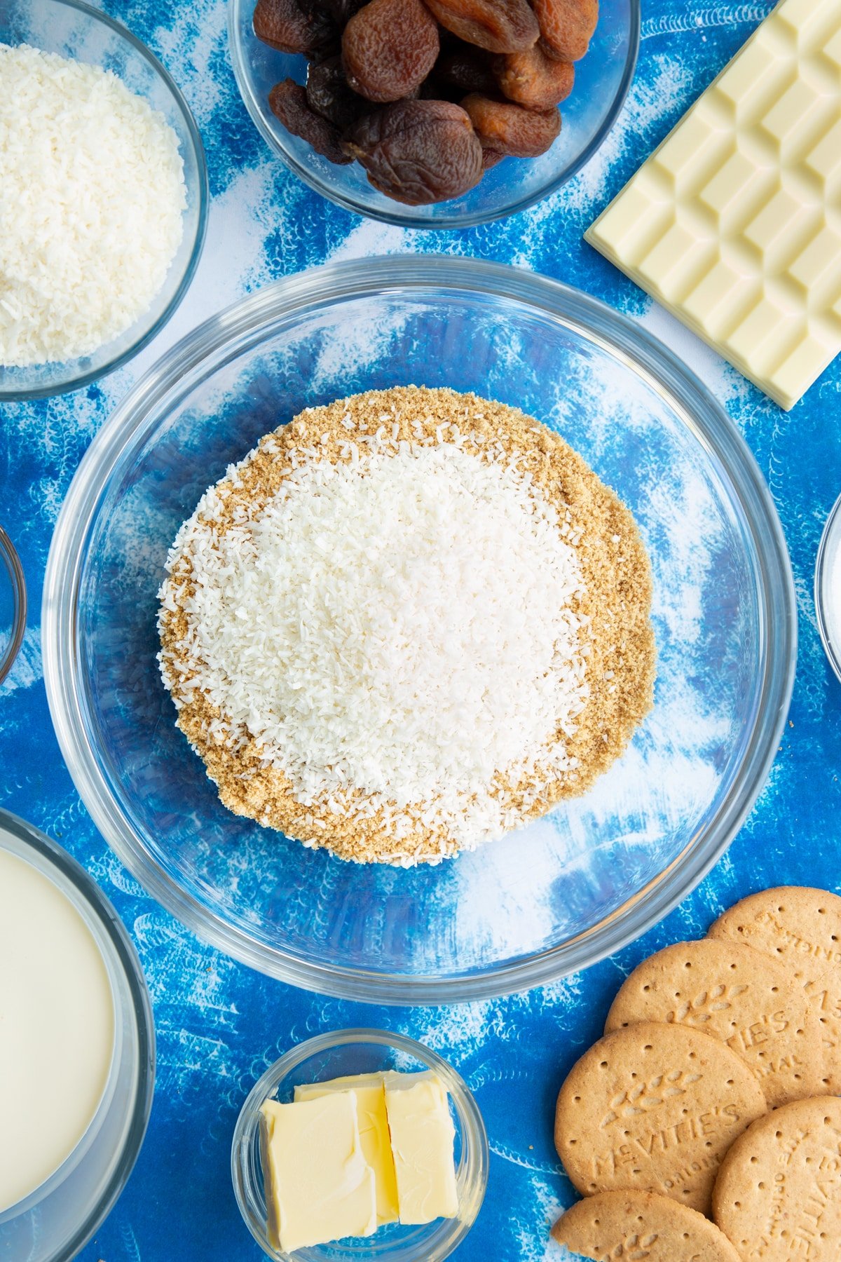 Crushed biscuits and desiccated coconut in a bowl. Ingredients to make an apricot cheesecake surround the bowl.