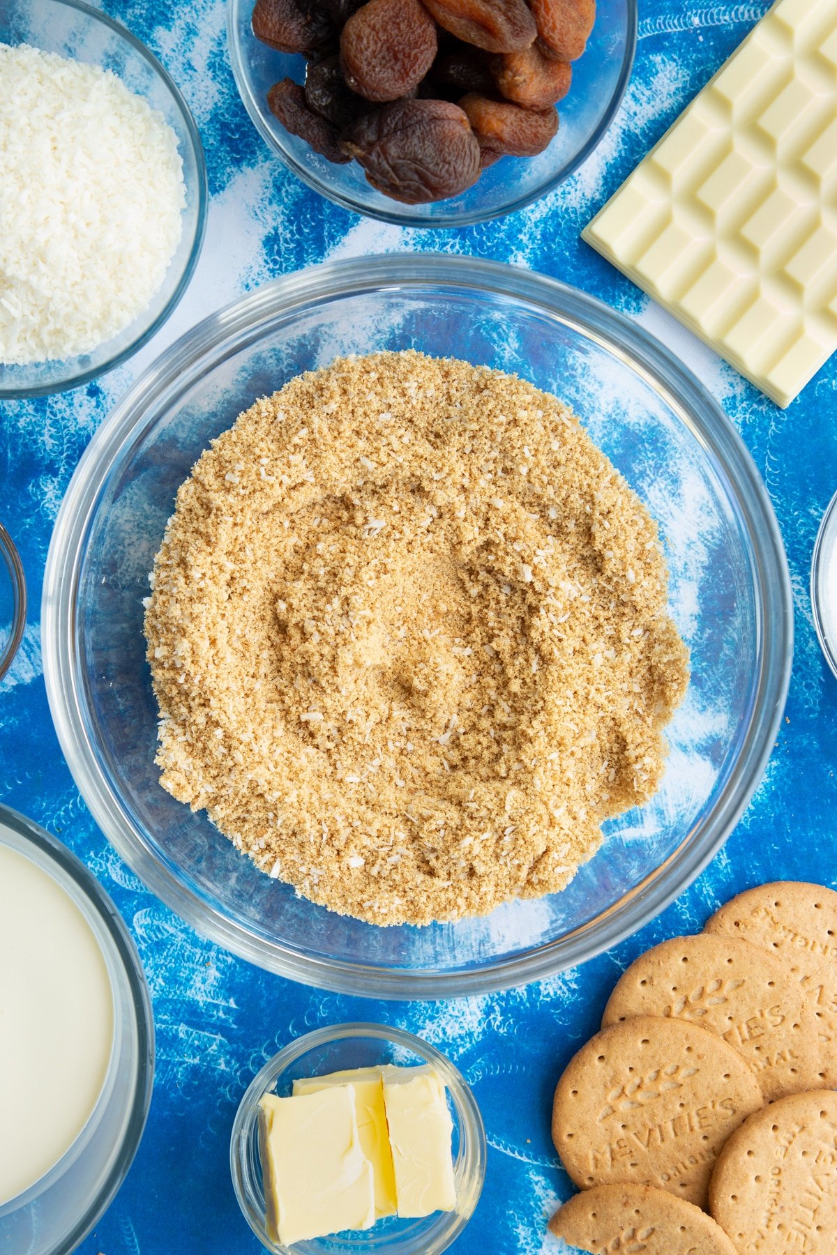 Crushed biscuits and desiccated coconut mixed together in a bowl. Ingredients to make an apricot cheesecake surround the bowl.