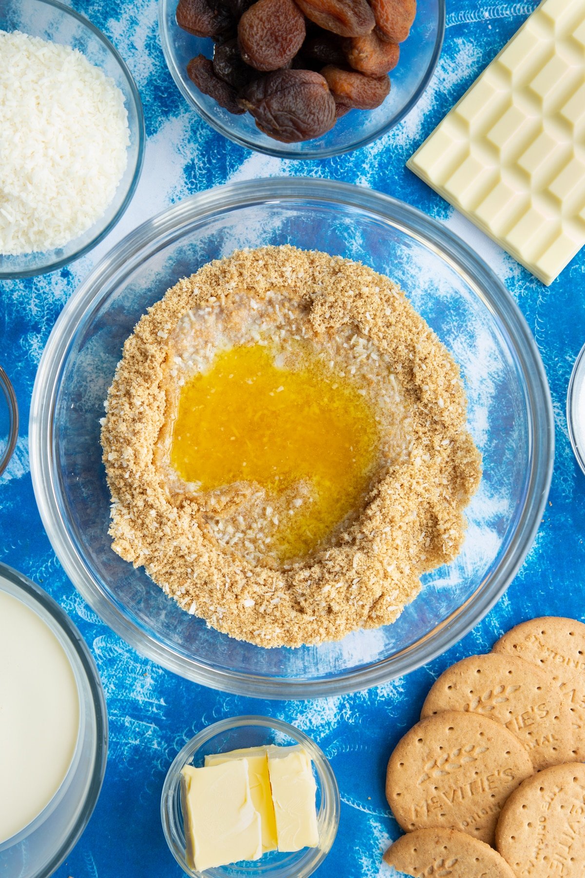 Crushed biscuits and desiccated coconut mixed together with melted butter on top in a bowl. Ingredients to make an apricot cheesecake surround the bowl.