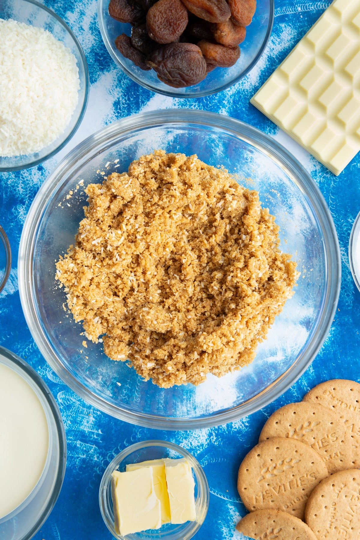 Crushed biscuits and desiccated coconut mixed together with melted butter in a bowl. Ingredients to make an apricot cheesecake surround the bowl.