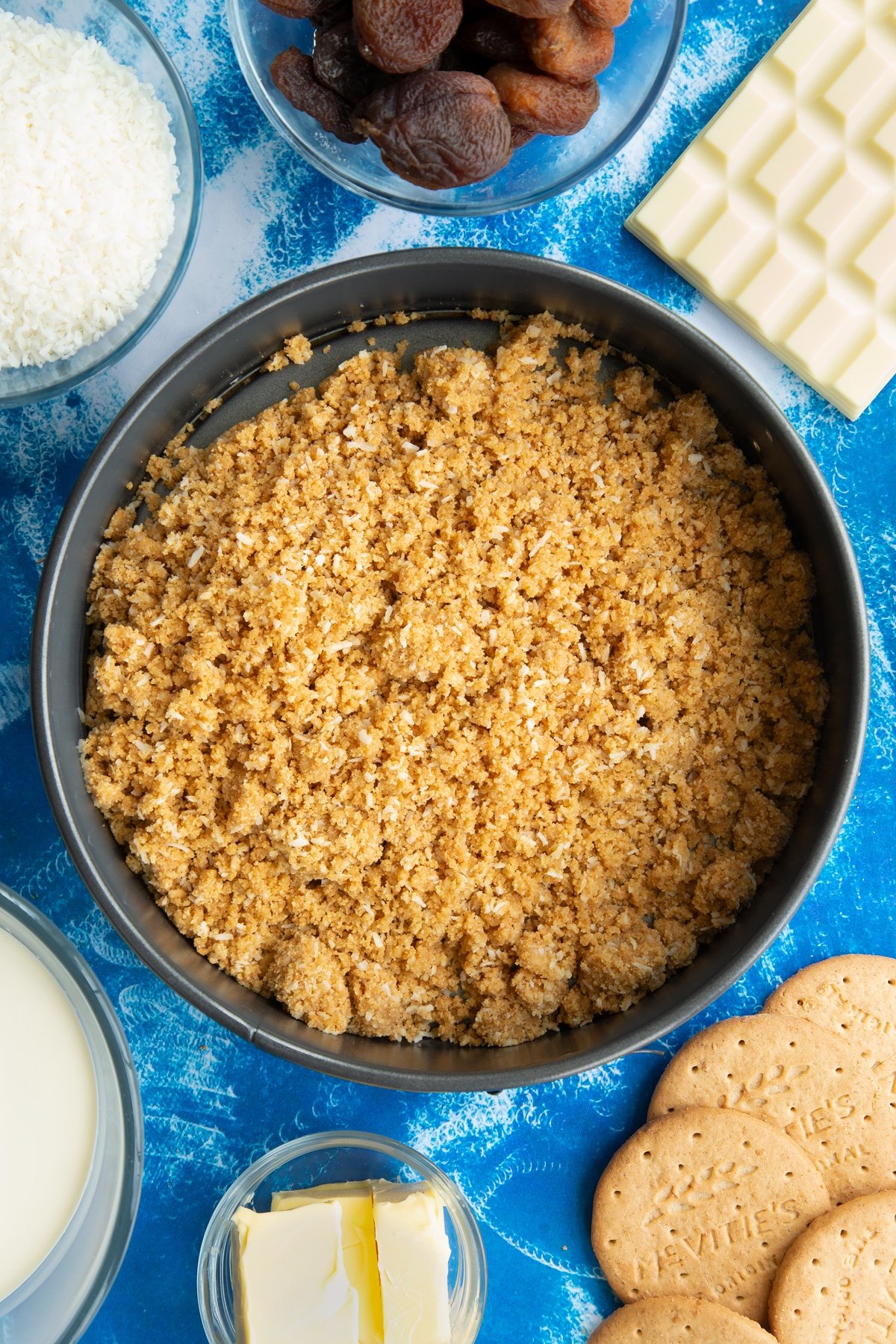 Crushed biscuits, coconut and butter in a tin. Ingredients to make an apricot cheesecake surround the tin.