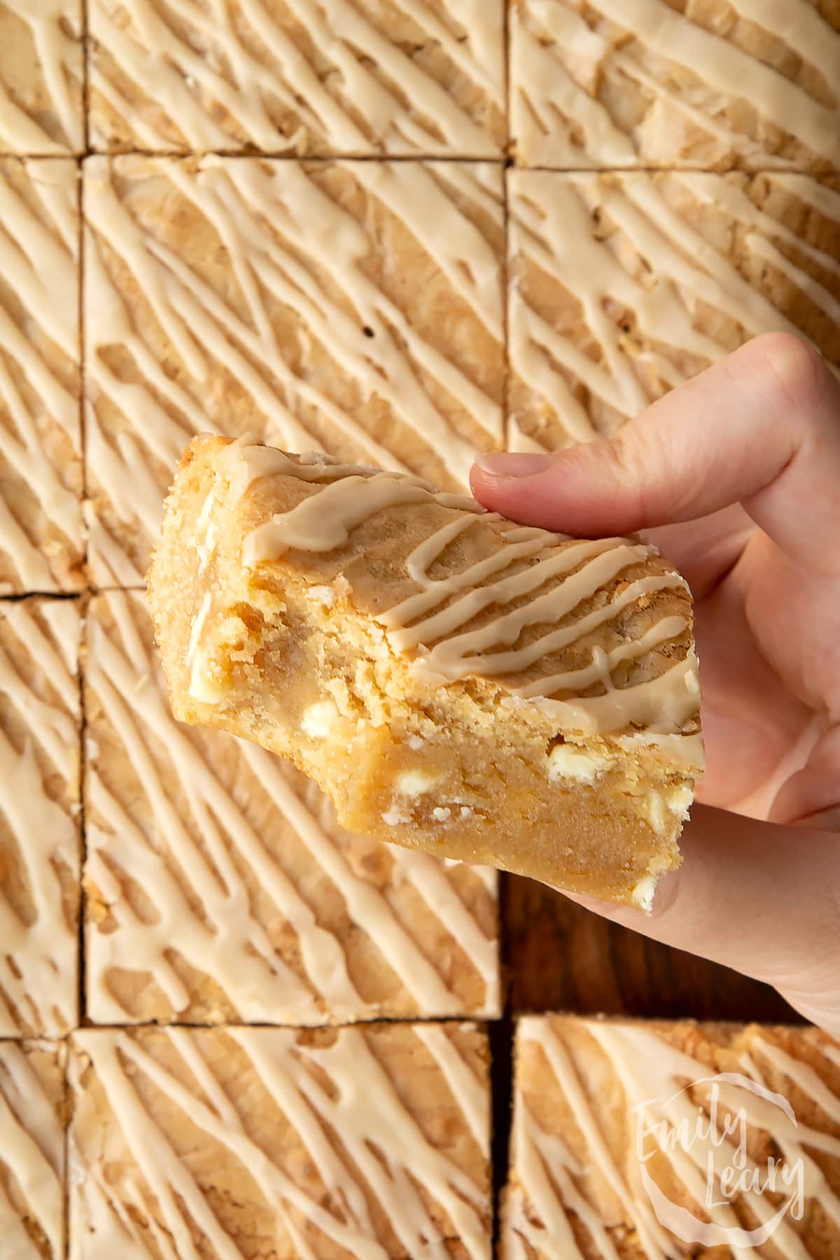 Hand holding a square of Baileys blondie with a bite out of it above more blondies.