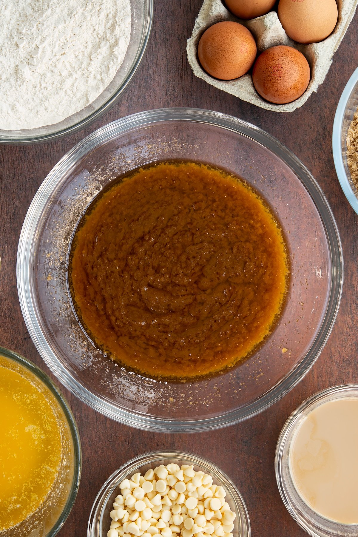 Melted butter and soft brown sugar whisked together in a glass mixing bowl. Ingredients to make Baileys blondies surround the bowl.