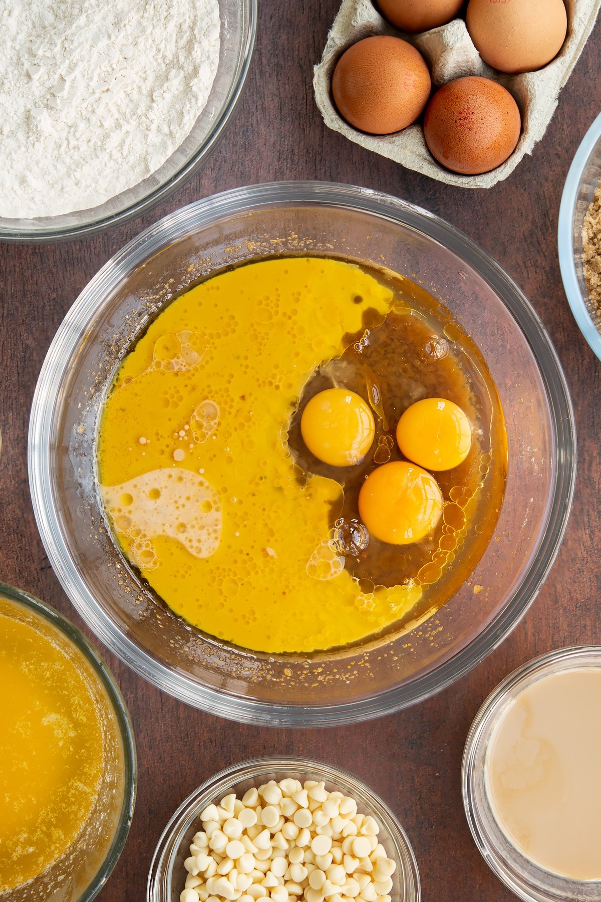 Melted butter, soft brown sugar, eggs and Baileys in a glass mixing bowl. Ingredients to make Baileys blondies surround the bowl.