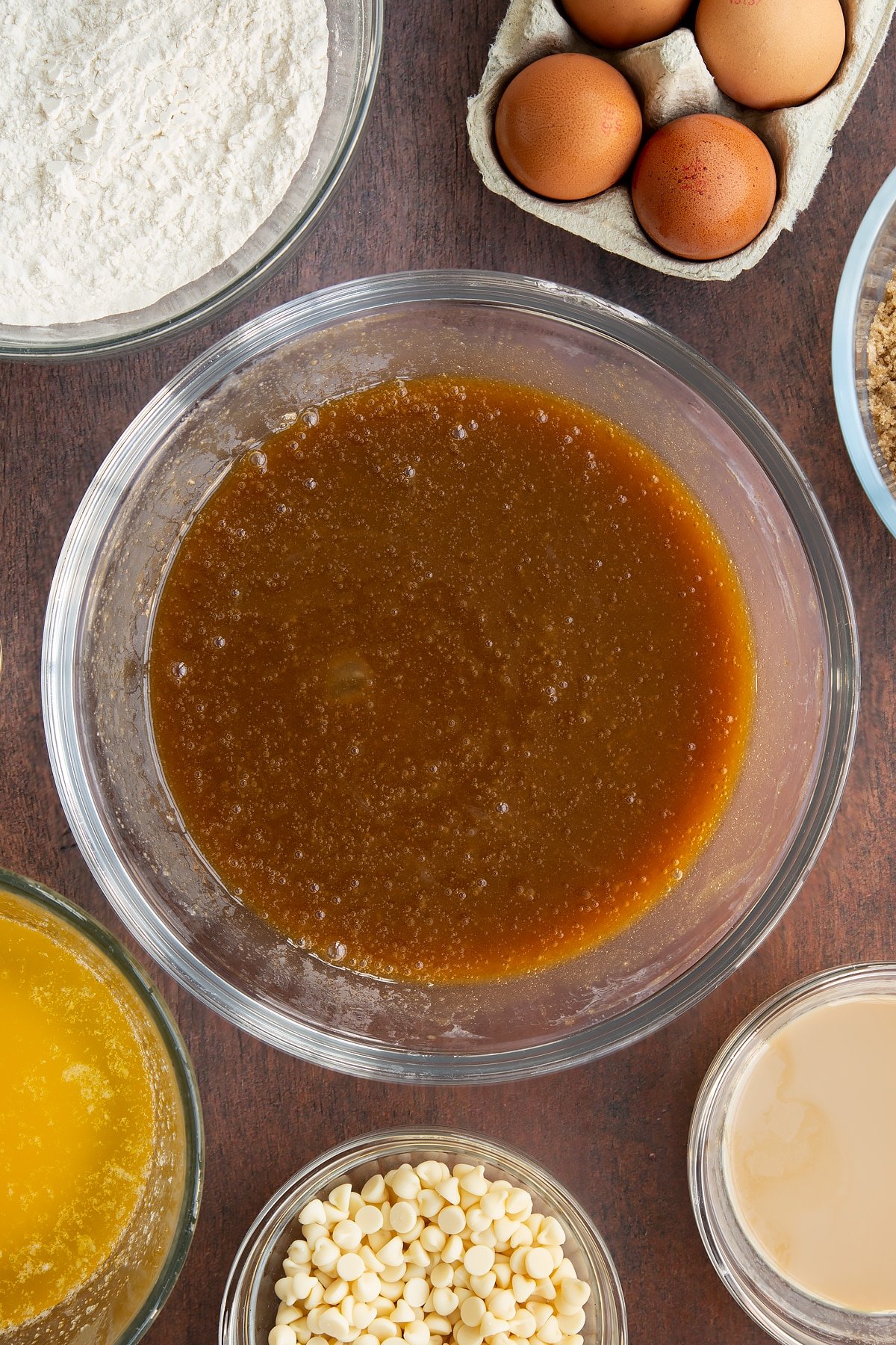 Melted butter, soft brown sugar, eggs and Baileys whisked together in a glass mixing bowl. Ingredients to make Baileys blondies surround the bowl.