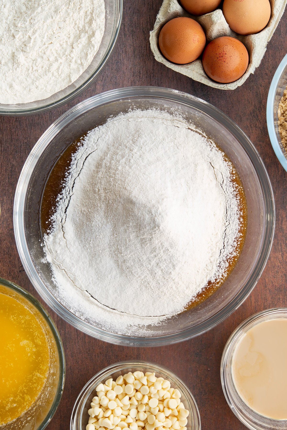 Melted butter, soft brown sugar, eggs and Baileys whisked together in a glass mixing bowl with flour and baking powder on top. Ingredients to make Baileys blondies surround the bowl.