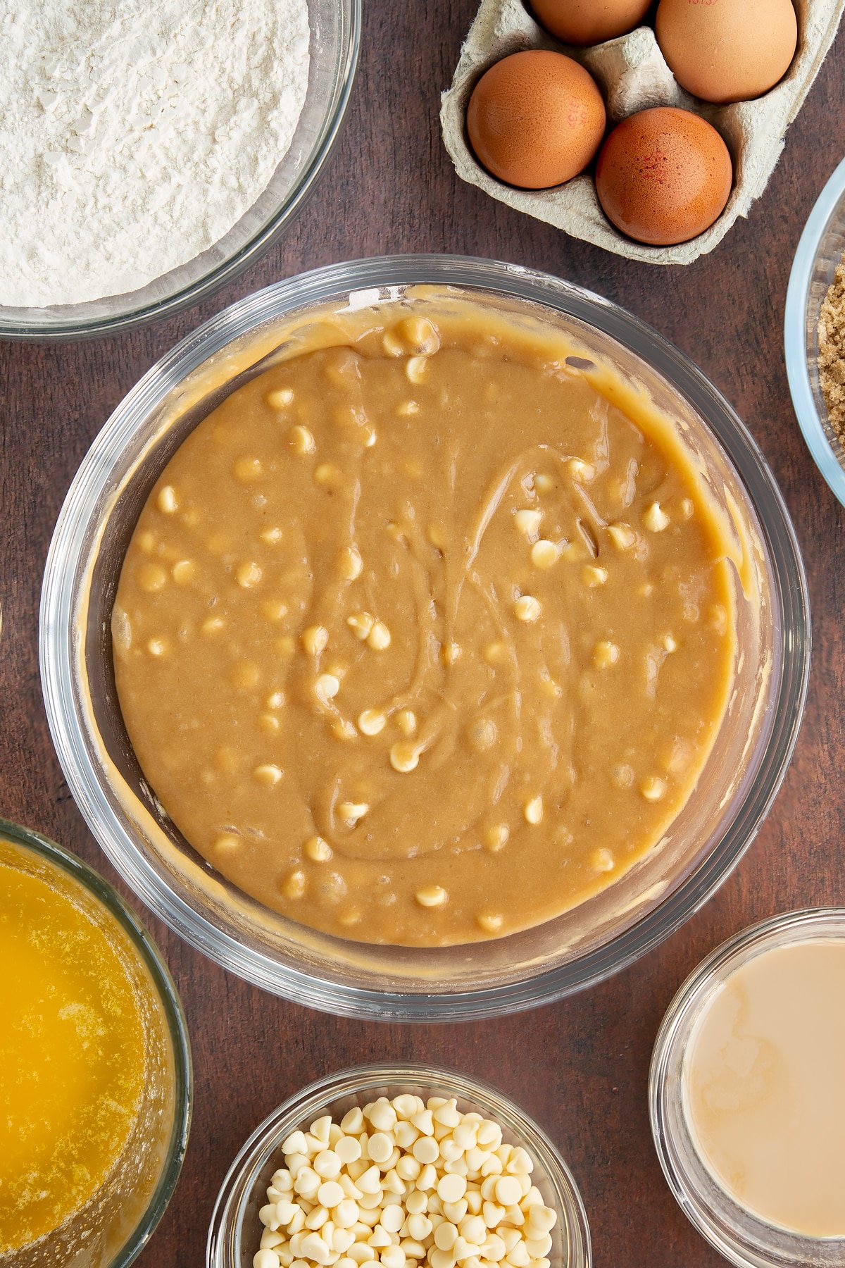 Baileys blondie batter with white chocolate chips in a glass mixing bowl. Ingredients to make Baileys blondies surround the bowl.