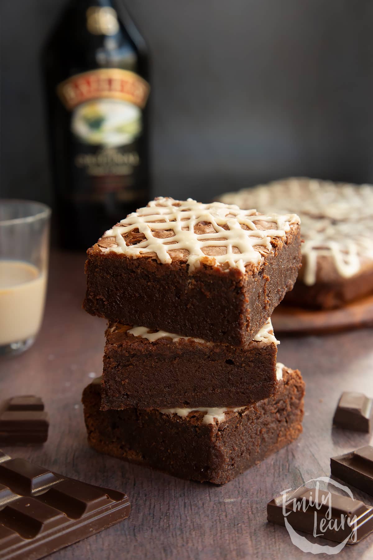 Stack of iced Baileys brownies, with more brownies and a bottle of Baileys in the background. 