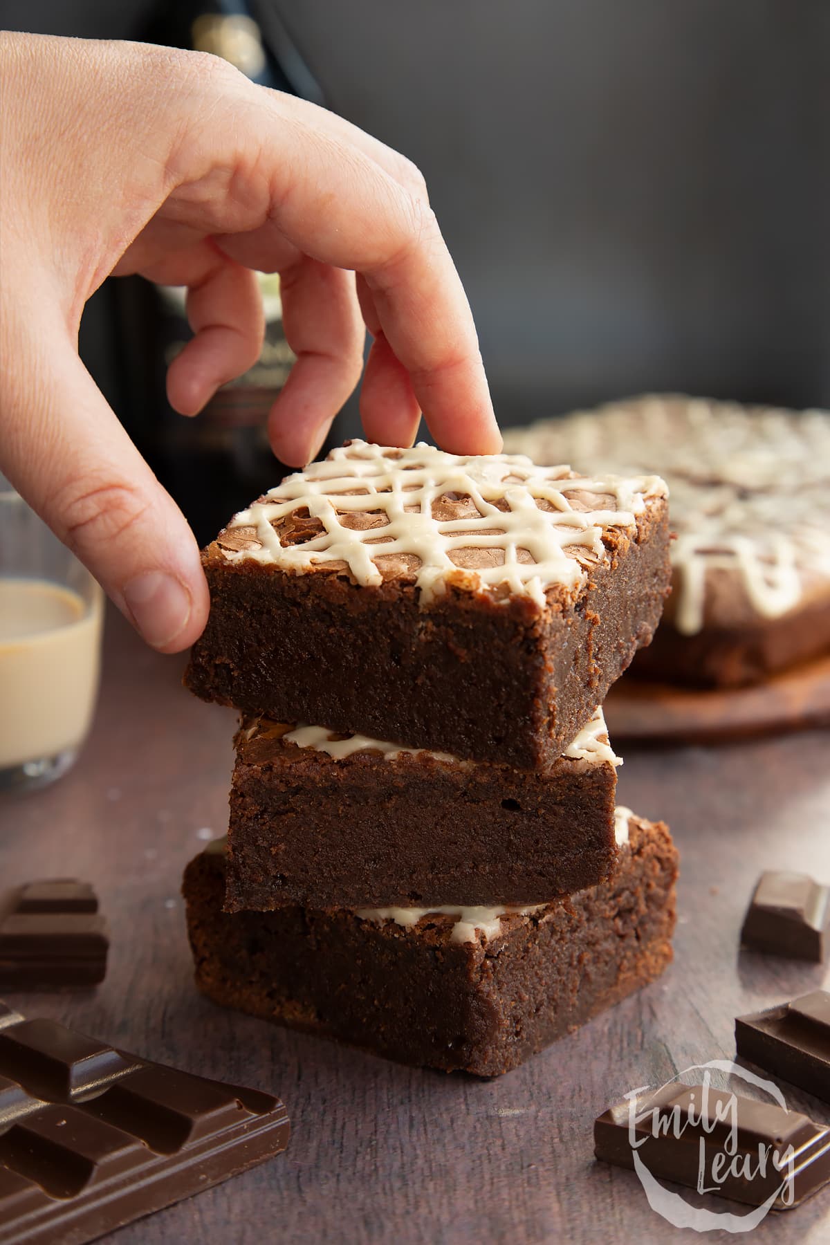 Stack of iced Baileys brownies, with more brownies and a bottle of Baileys in the background. A hand reaches for one.