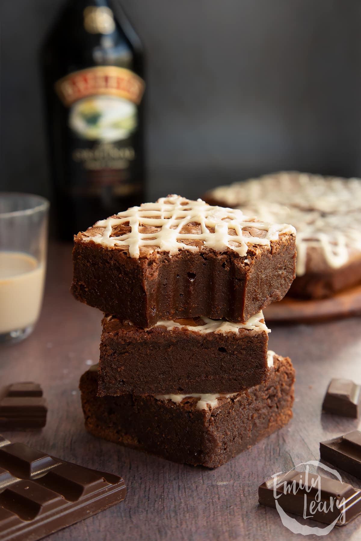 Stack of iced Baileys brownies, with more brownies and a bottle of Baileys in the background. One brownie has a bite out of it.