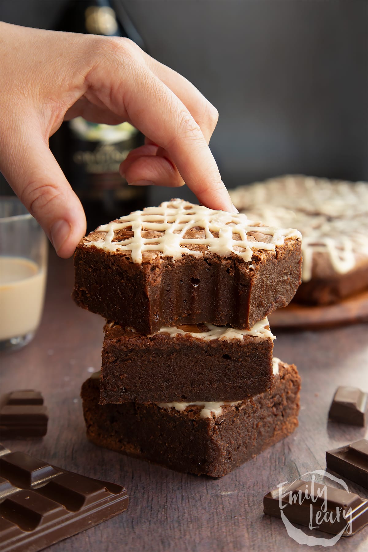 Stack of iced Baileys brownies, with more brownies and a bottle of Baileys in the background. One has a bite out if and a hand reaches for it.