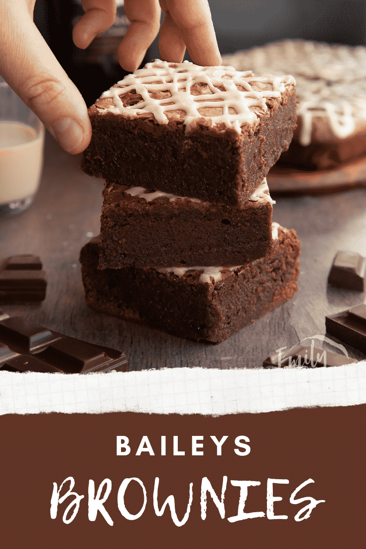 Stack of iced Baileys brownies, with more brownies and a bottle of Baileys in the background. A hand reaches for one. Caption reads: Baileys brownies.