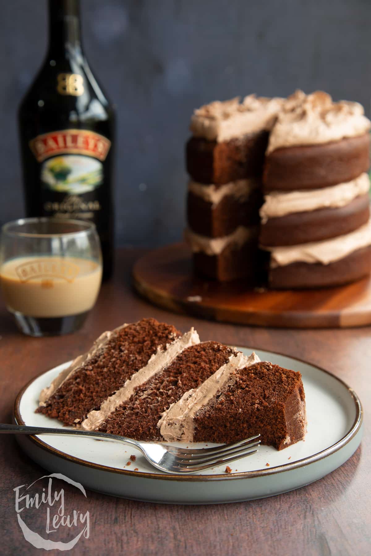 Slice of chocolate Baileys cake with Baileys buttercream frosting on a white plate with a piece cut away and a fork resting beside it.