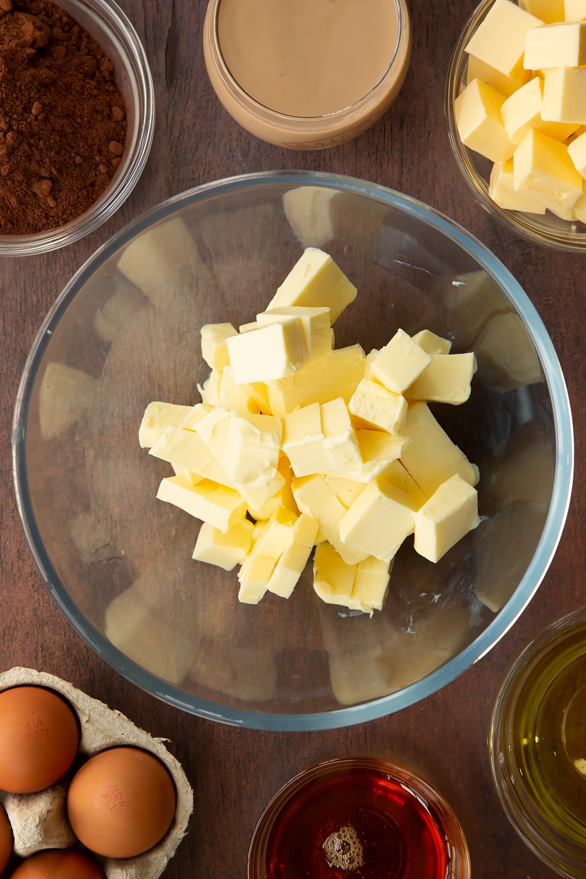 Cubed butter in a glass mixing bowl. Ingredients to make a Baileys cake surround the bowl.