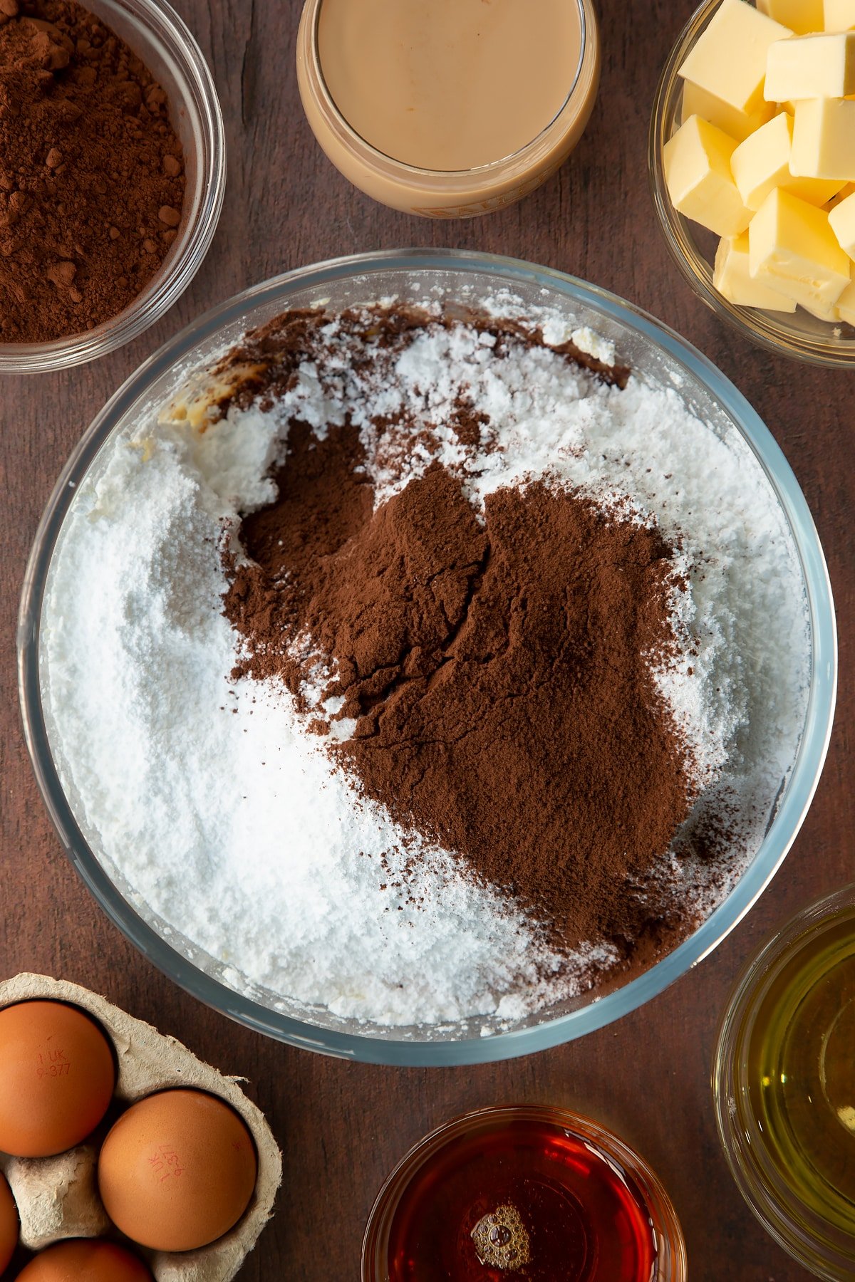 Whipped butter in a glass mixing bowl, topped with icing sugar, cocoa and Baileys. Ingredients to make a Baileys cake surround the bowl.