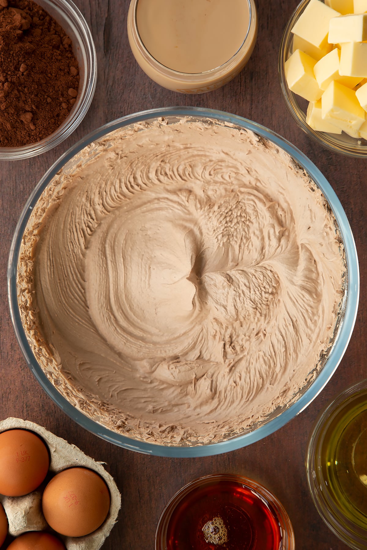 Baileys buttercream icing in a glass mixing bowl. Ingredients to make a Baileys cake surround the bowl.